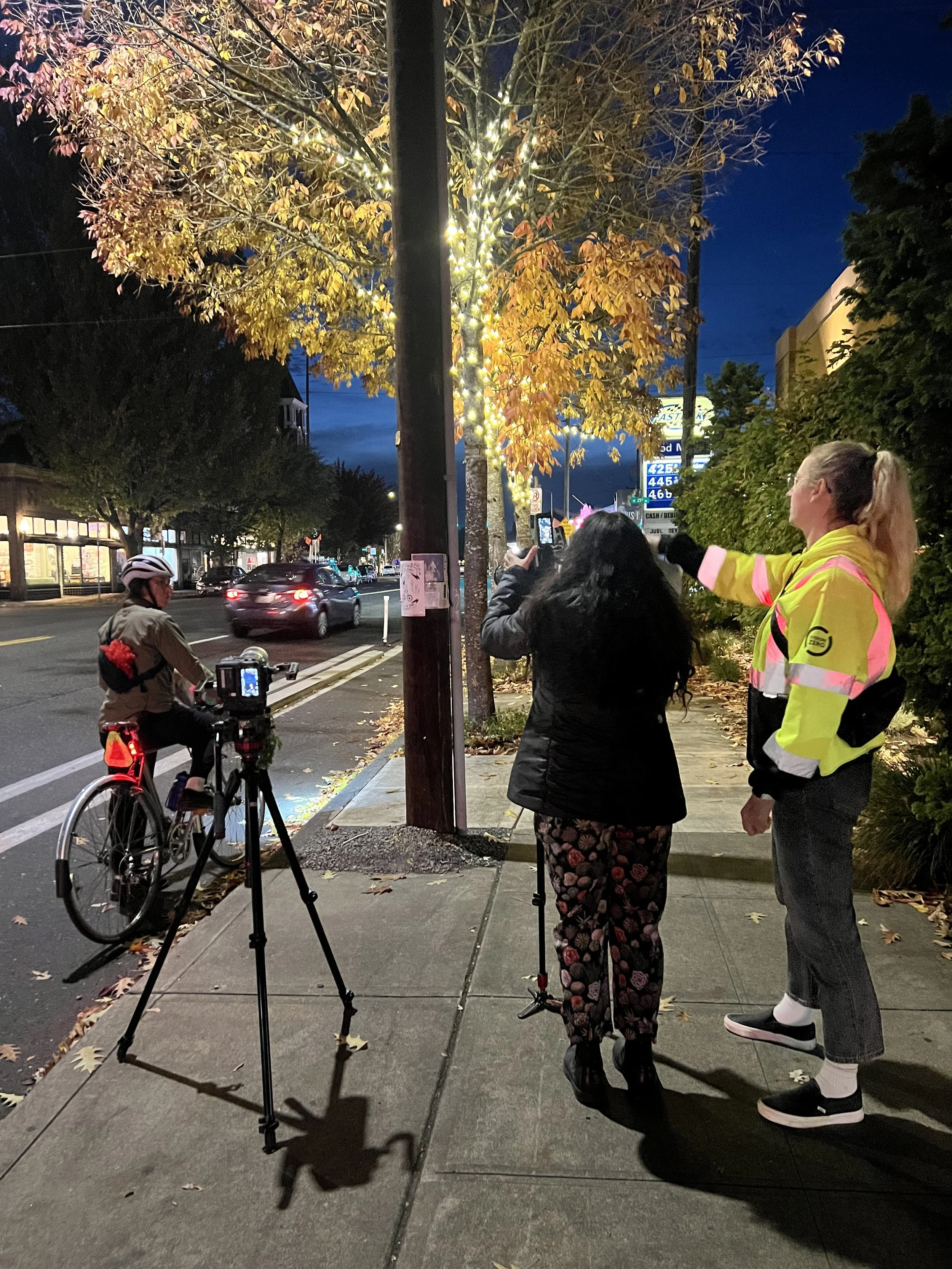 A street scene at dusk with a tree decorated with string lights. People are gathered on the sidewalk, some taking photos. A cyclist is nearby, and cars are driving along the street. The sky is dark, and there are shops and traffic signs visible in the background.