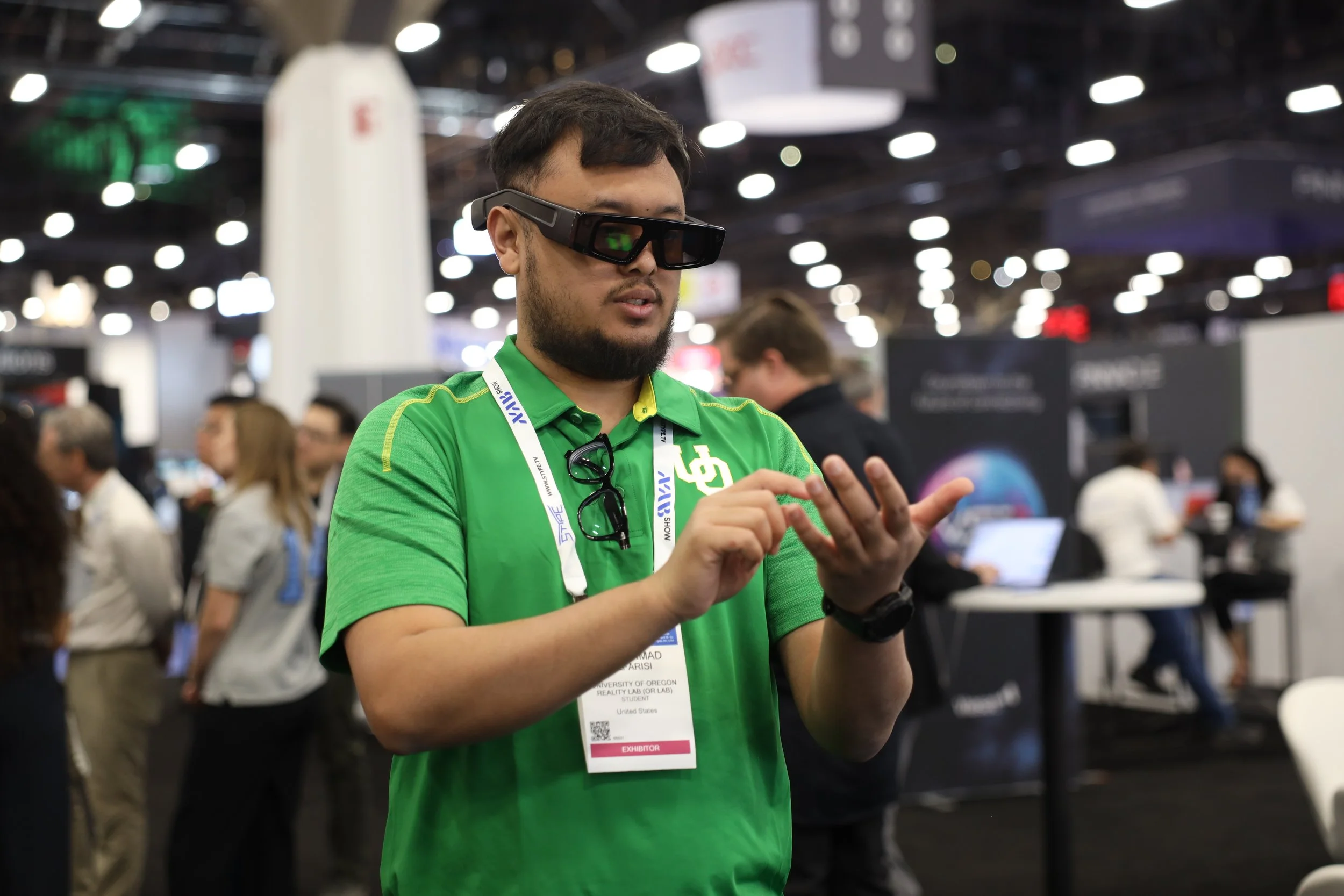 A man in a green shirt with a name badge and conference lanyard is wearing mixed reality glasses and interacting with a virtual device at a busy technology event.