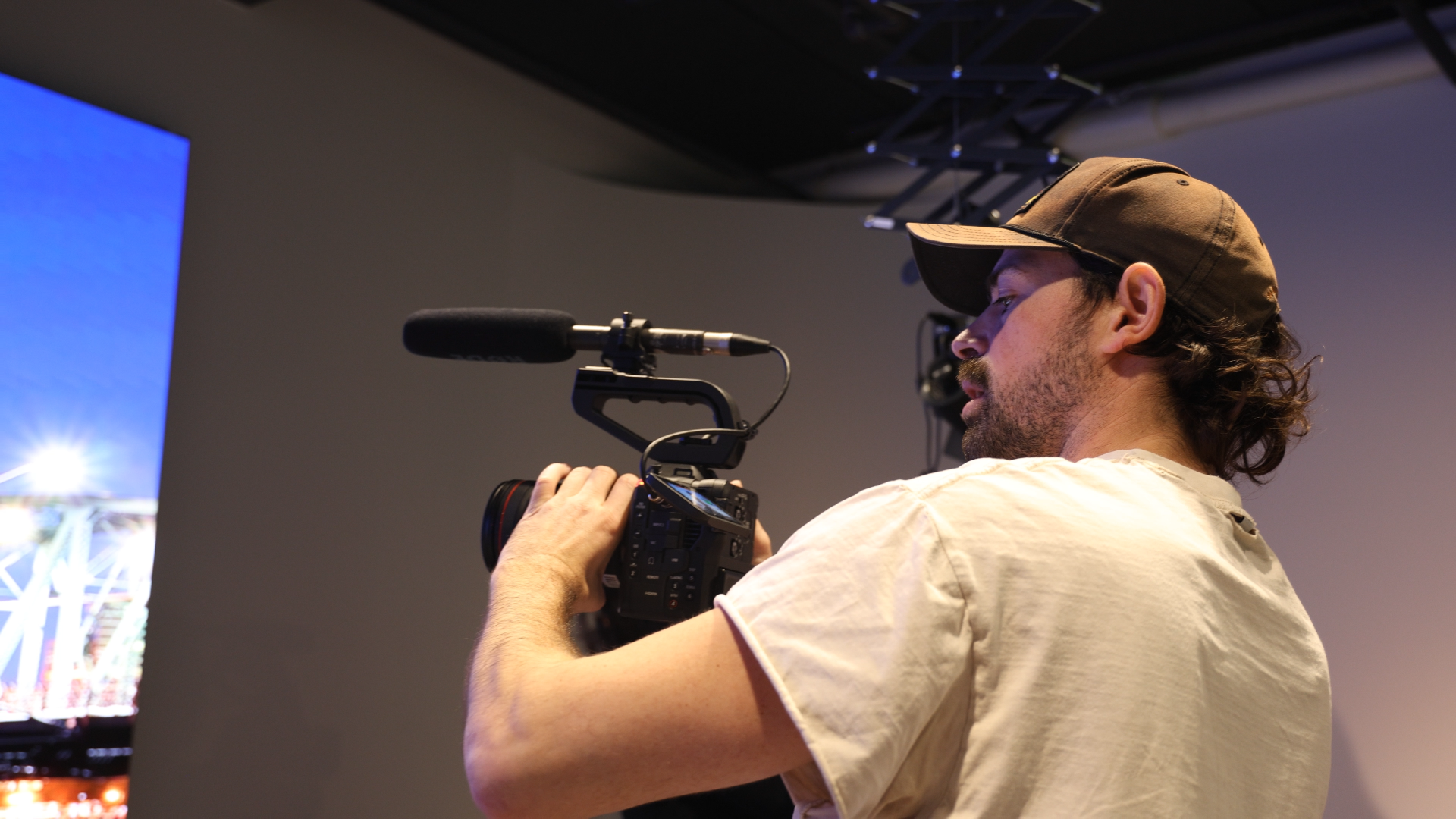Man filming with professional camera in an indoor setting with a large screen displaying a bright colorful image.