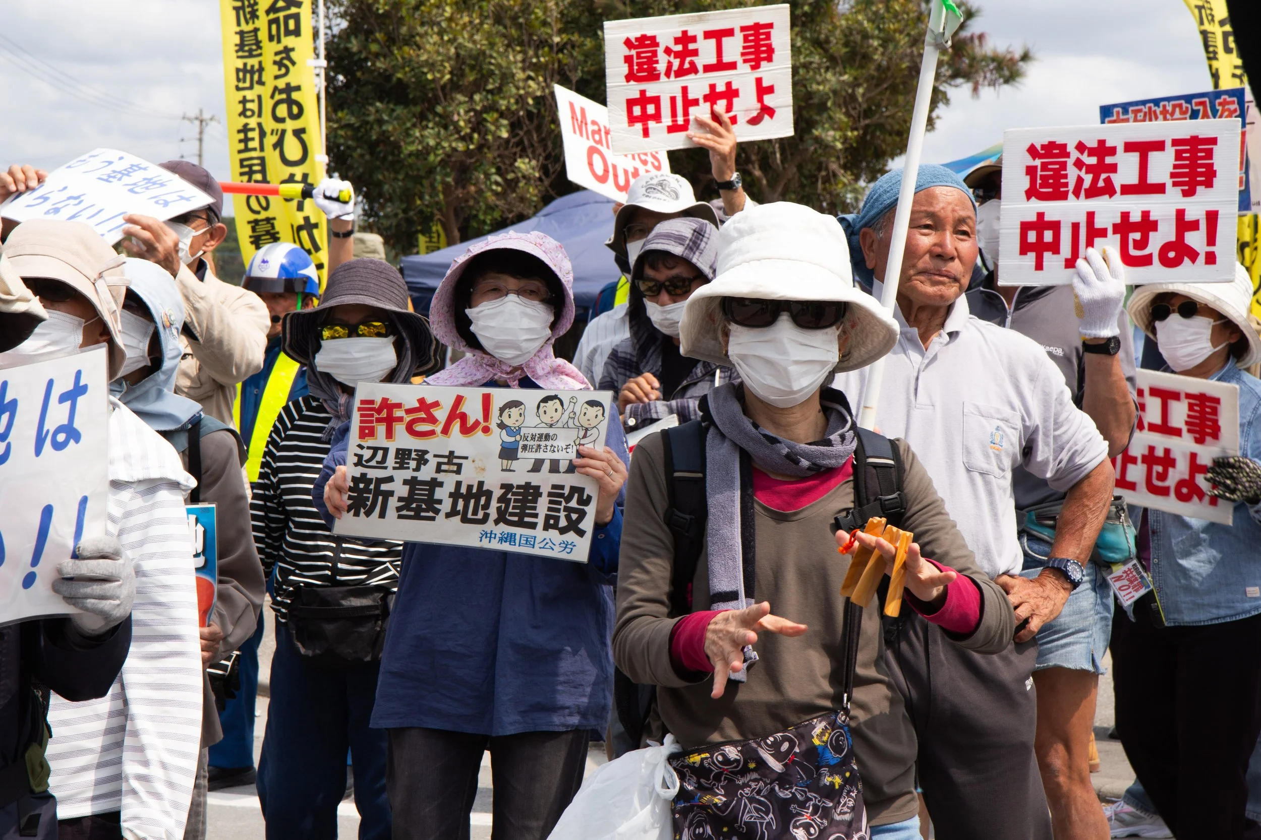 A group of protesters, mostly elderly, wearing face masks and hats, holding signs with Japanese and some English text, protesting against illegal construction. Yellow and white banners are visible among them.