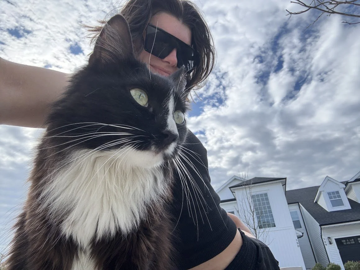 A person wearing sunglasses and a black shirt is posing with a black and white cat outdoors on a cloudy day. The person's hair is partially covering their face, and they are smiling.