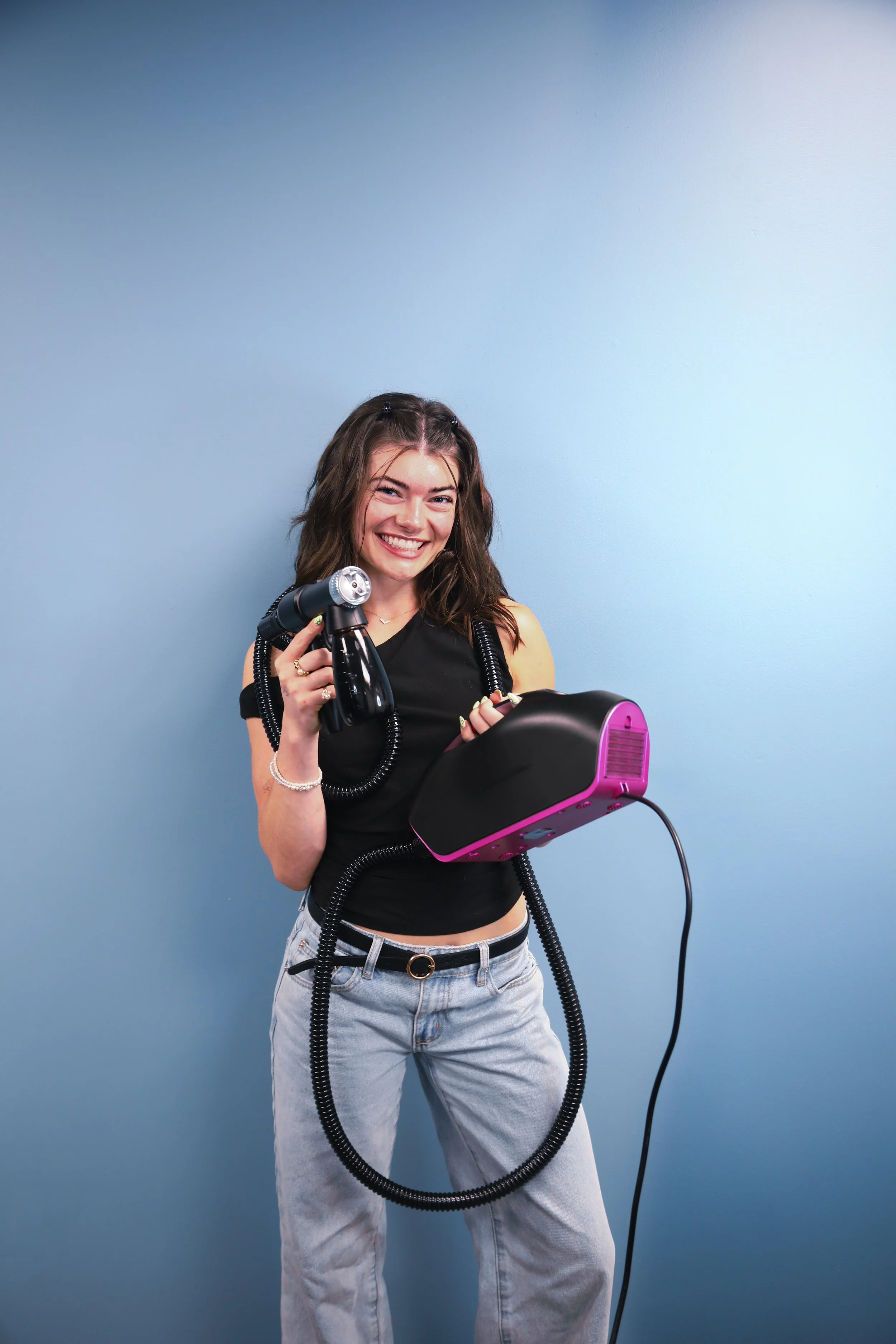 black off the shoulder top, jeans, holding spray tan machine and smiling in woodbury