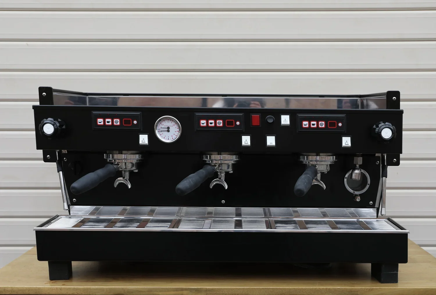 Commercial black espresso machine with three group heads, gauges, and control switches, placed on a wooden countertop in front of a beige garage door.