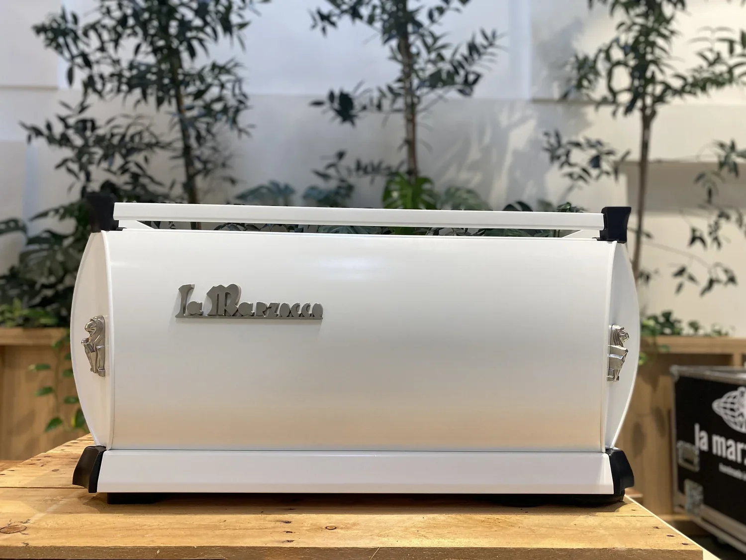 A white La Marzocco espresso machine on a wooden counter, with a blurred green plant in the background.