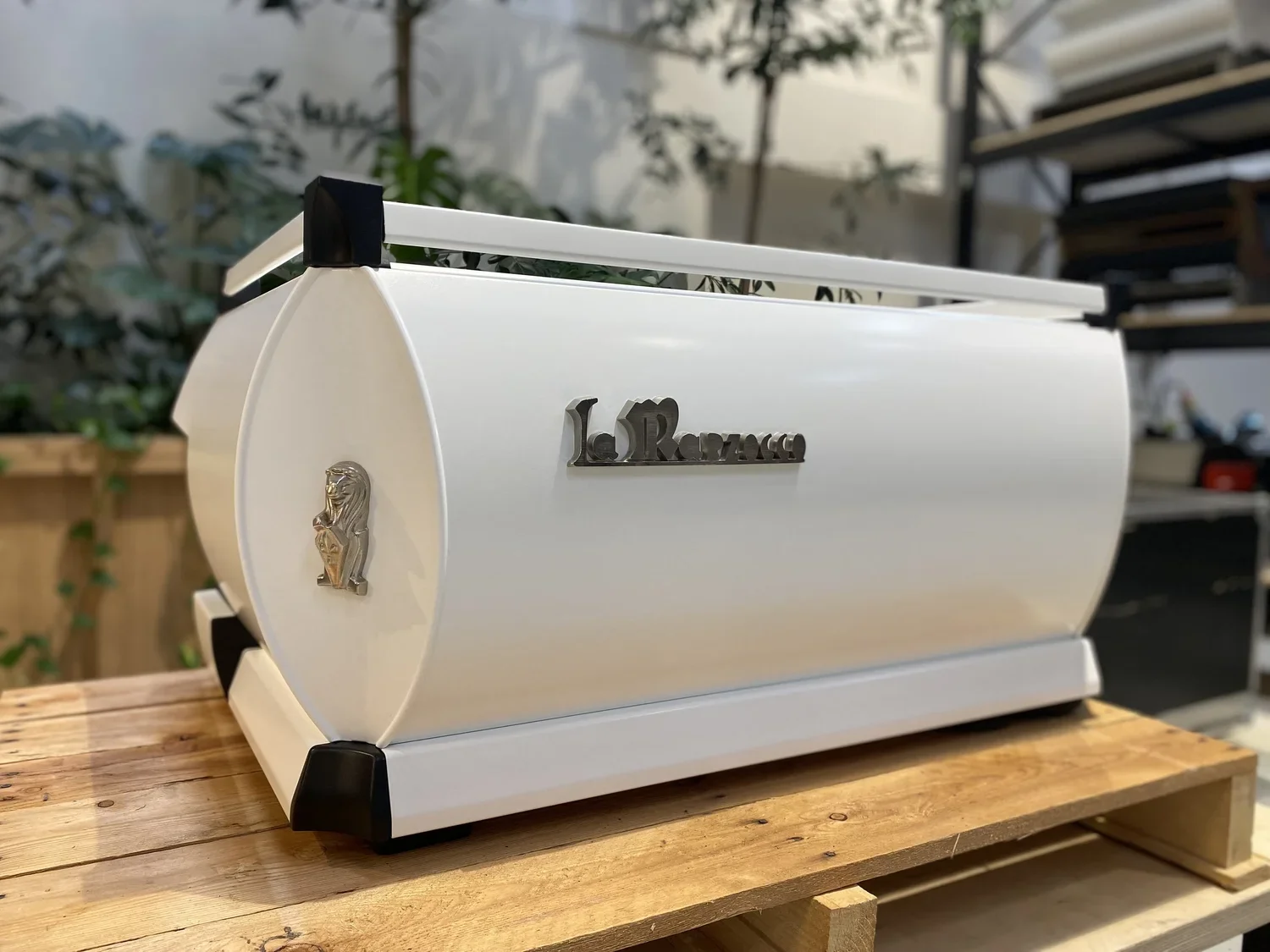 A white La Marzocco espresso machine with black accents, placed on a wooden surface, with a lion emblem on the front, in an outdoor setting with plants and shelves in the background.
