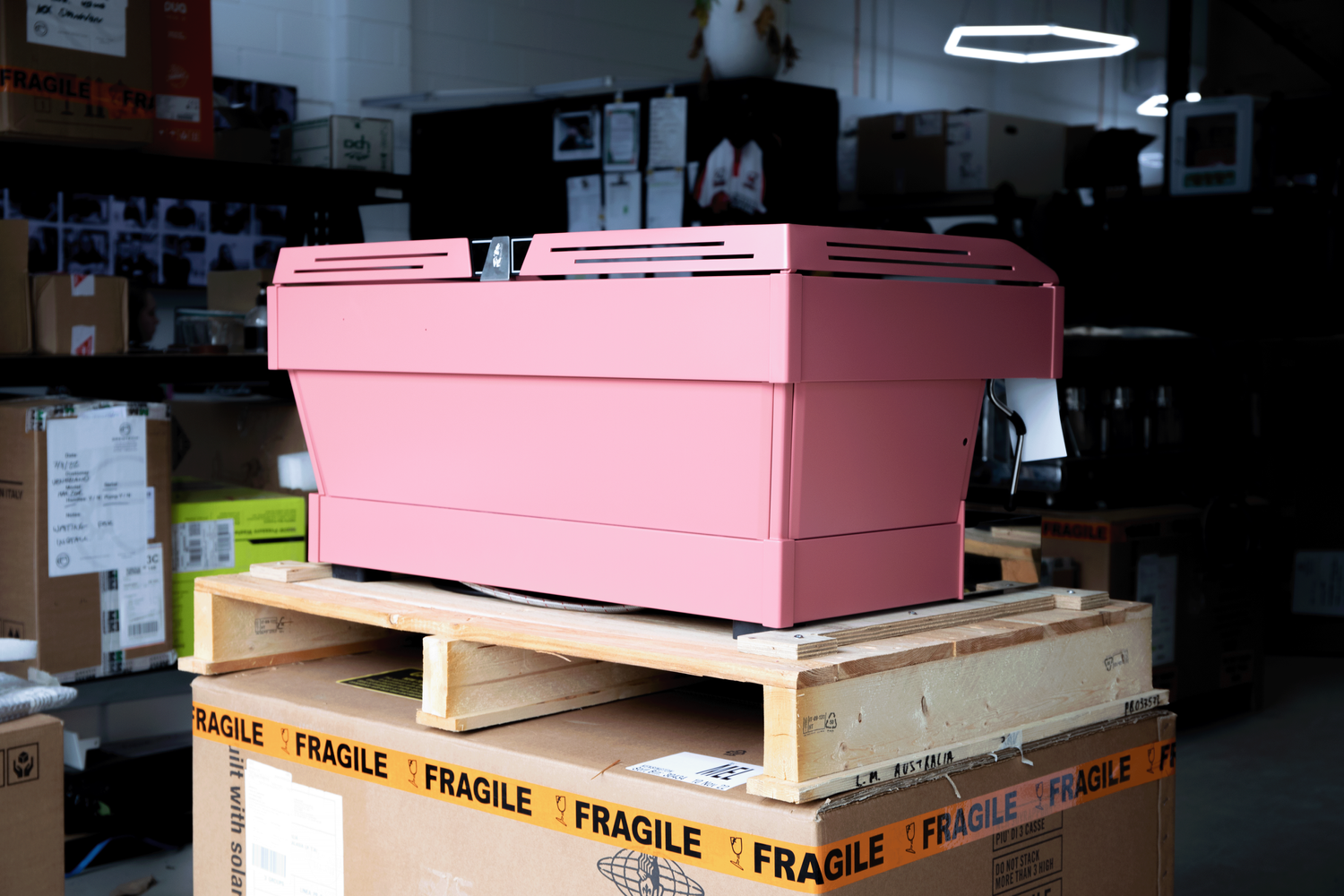 Pink printer on a wooden pallet in a warehouse or storage room with boxes and shelves in the background.