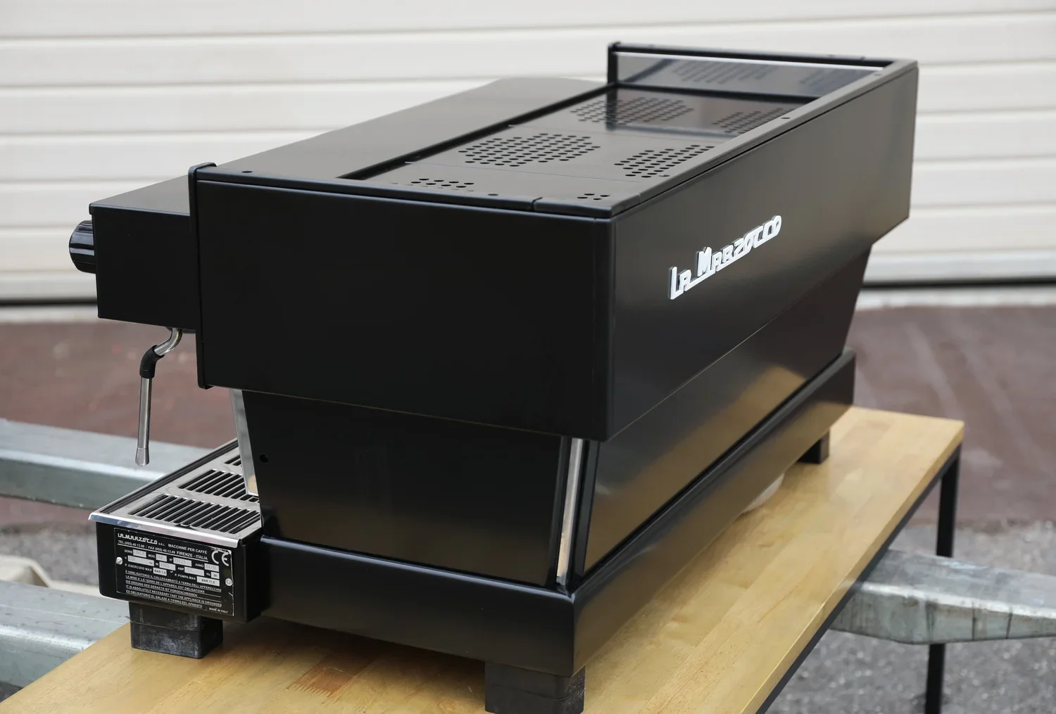 A black La Marzocco commercial espresso machine placed on a wooden table outdoors, with a metal roller shutter in the background.