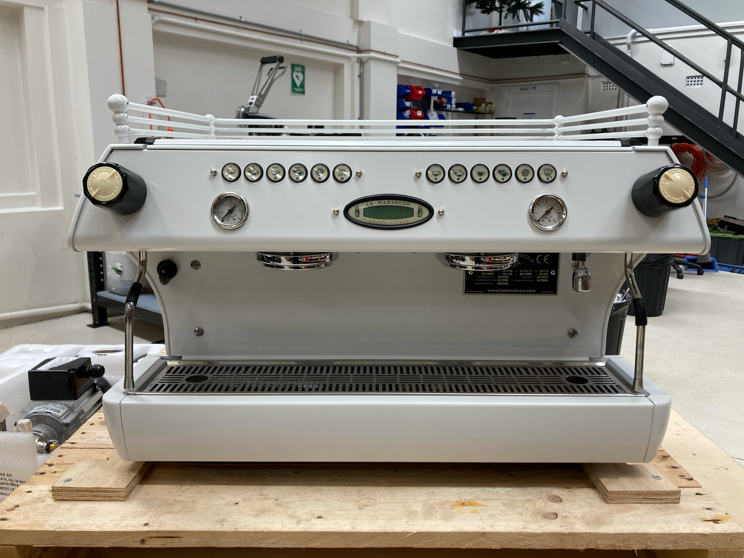 A white commercial espresso machine with control buttons, two pressure gauges, and large cream-colored knobs on either side, placed on a wooden table in a workshop setting.