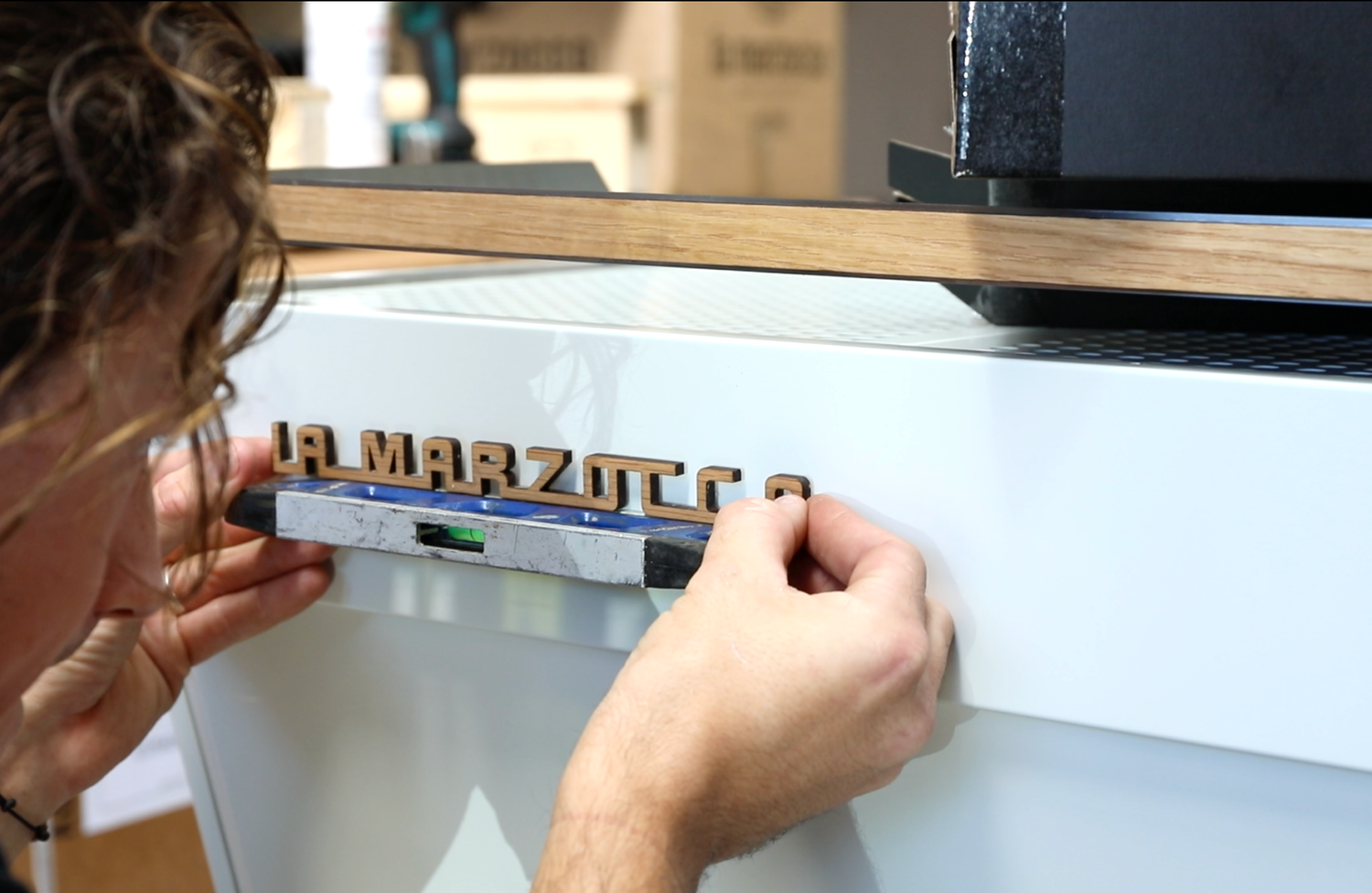 Person attaching wooden letters spelling 'LA MARZOCCO' to a machine or appliance.