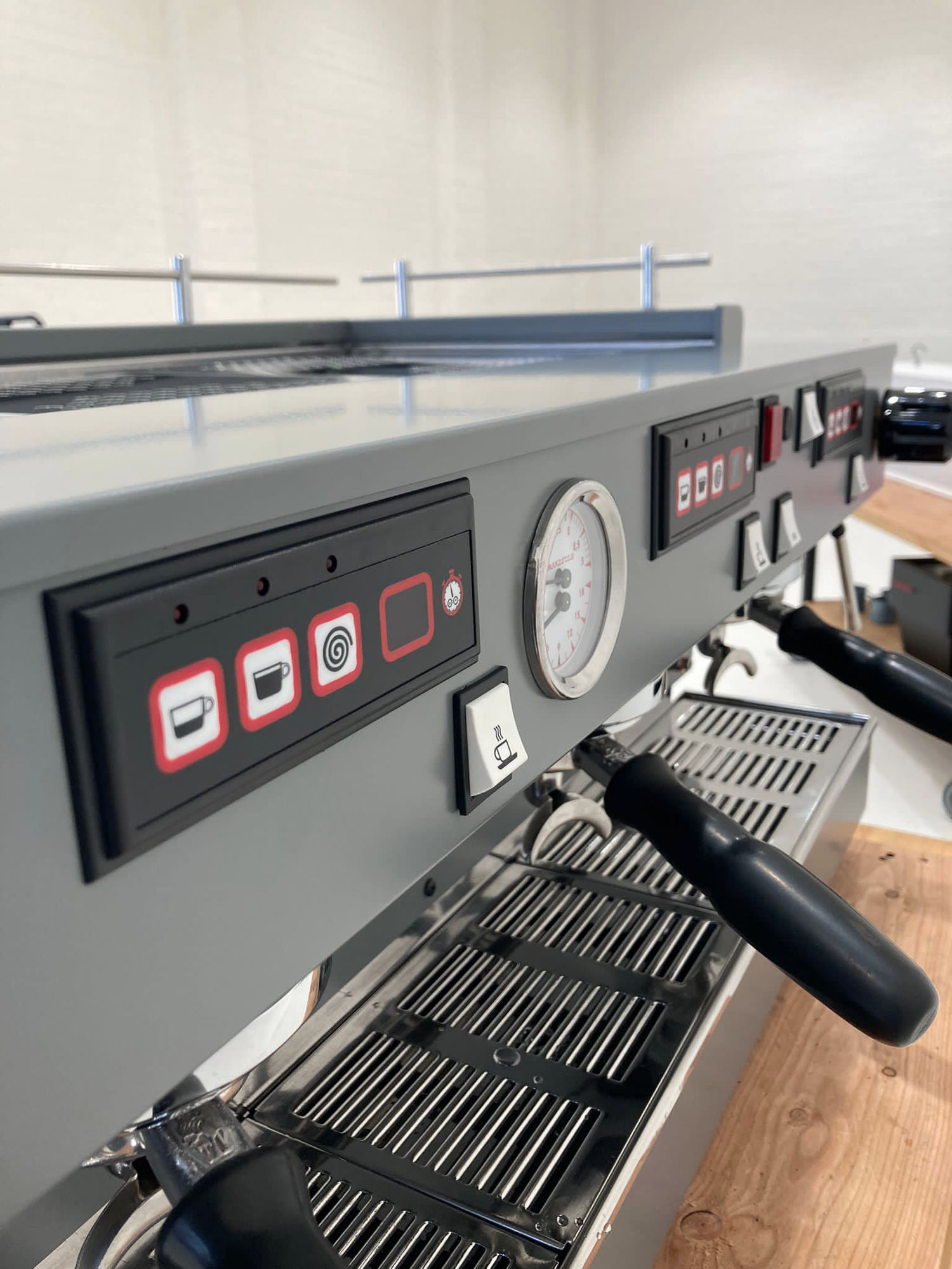 Close-up of a commercial espresso machine with a milk frothing steam wand, control panel, and a pressure gauge, positioned on a wooden countertop.