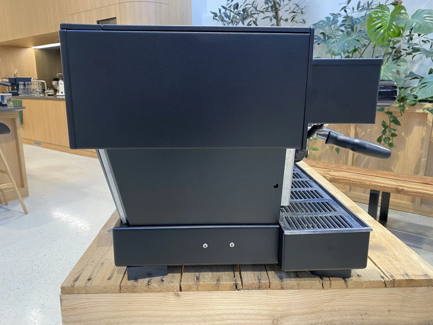 Black commercial espresso machine on a wooden table in a cafe with plants and wooden furniture in the background.