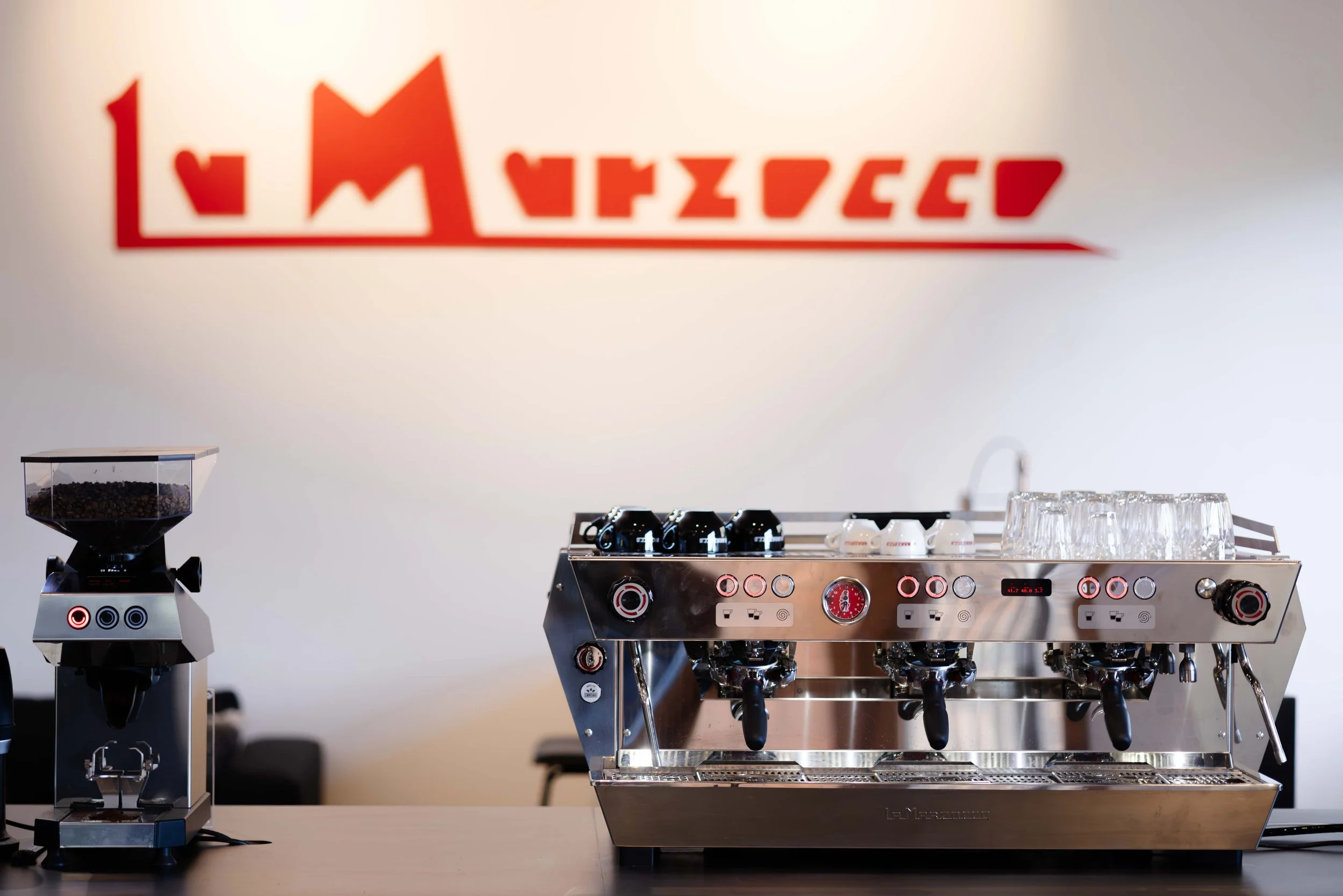 A modern espresso machine with a display and multiple handles, alongside a coffee grinder filled with coffee beans, set on a table in a minimalist setting.