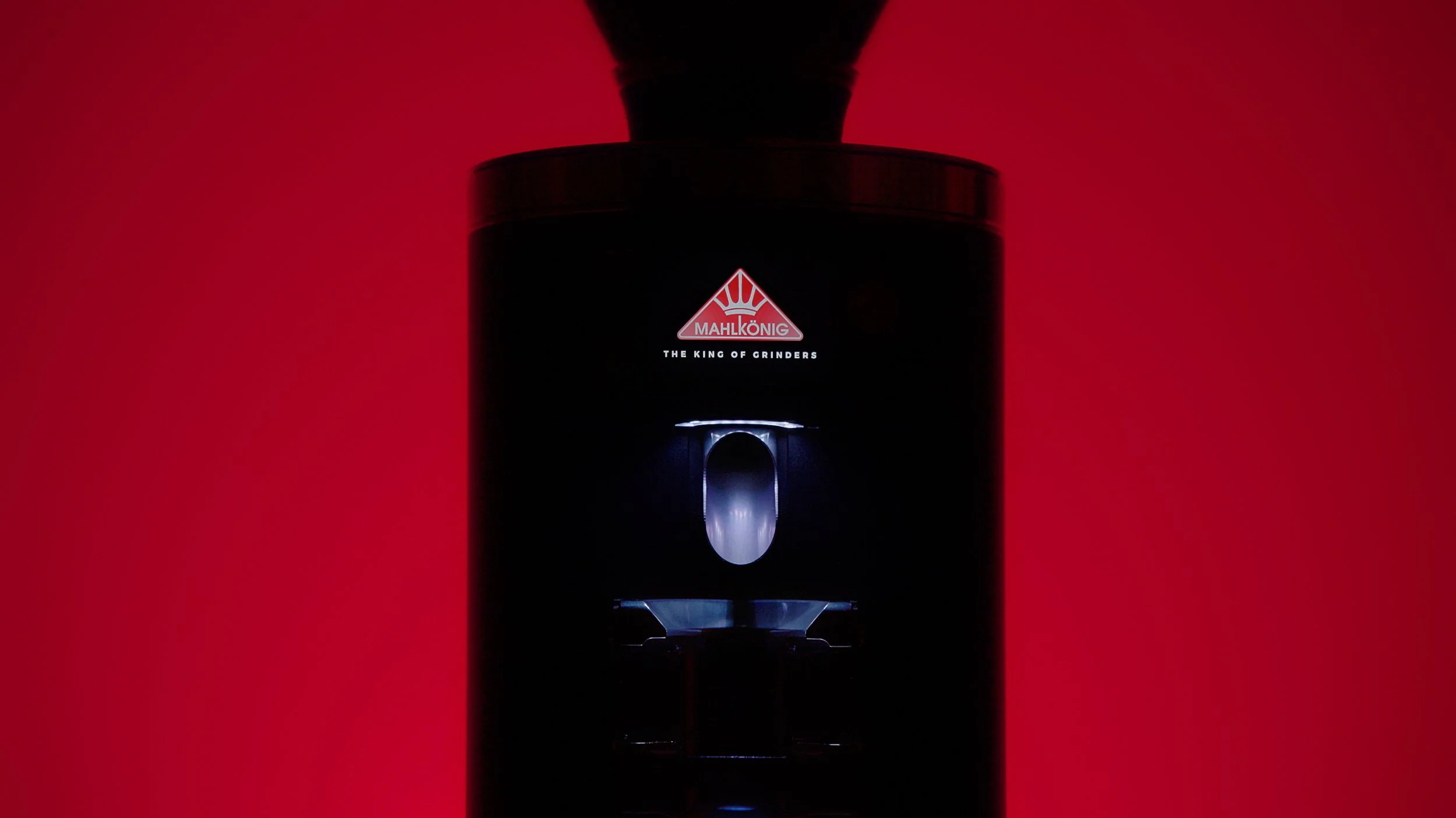 Close-up of a black Mahkönig coffee grinder with a red background, logo and tagline visible, illuminated from below.