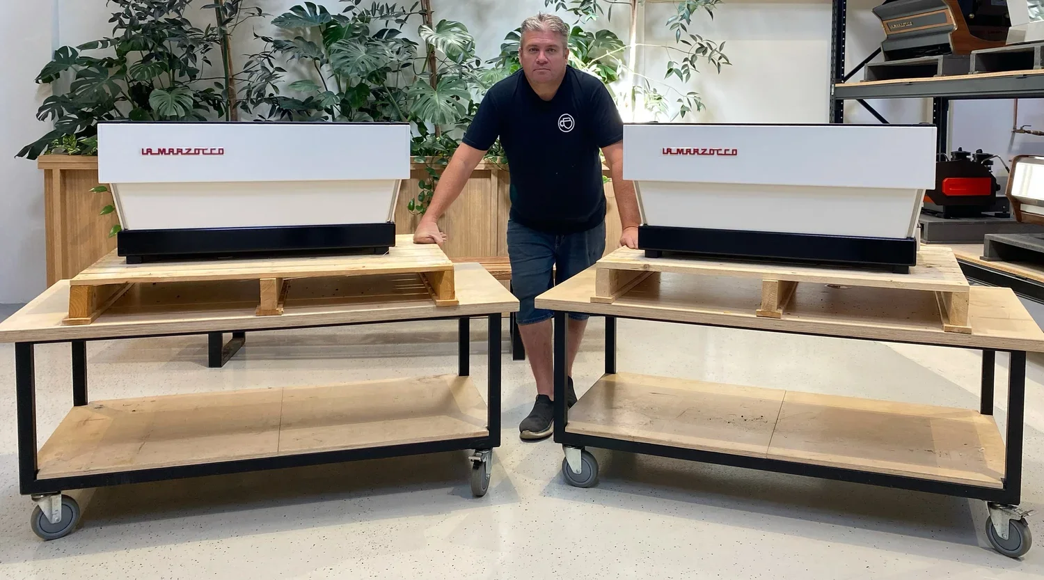 A Brewtech technician standing between two large white printers on wooden platforms on wheels in a room with plants and shelving.