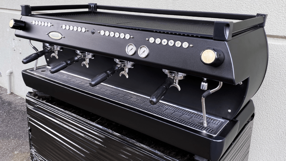 Black commercial espresso machine with multiple levers, knobs, and gauges, set against a light-colored wall.