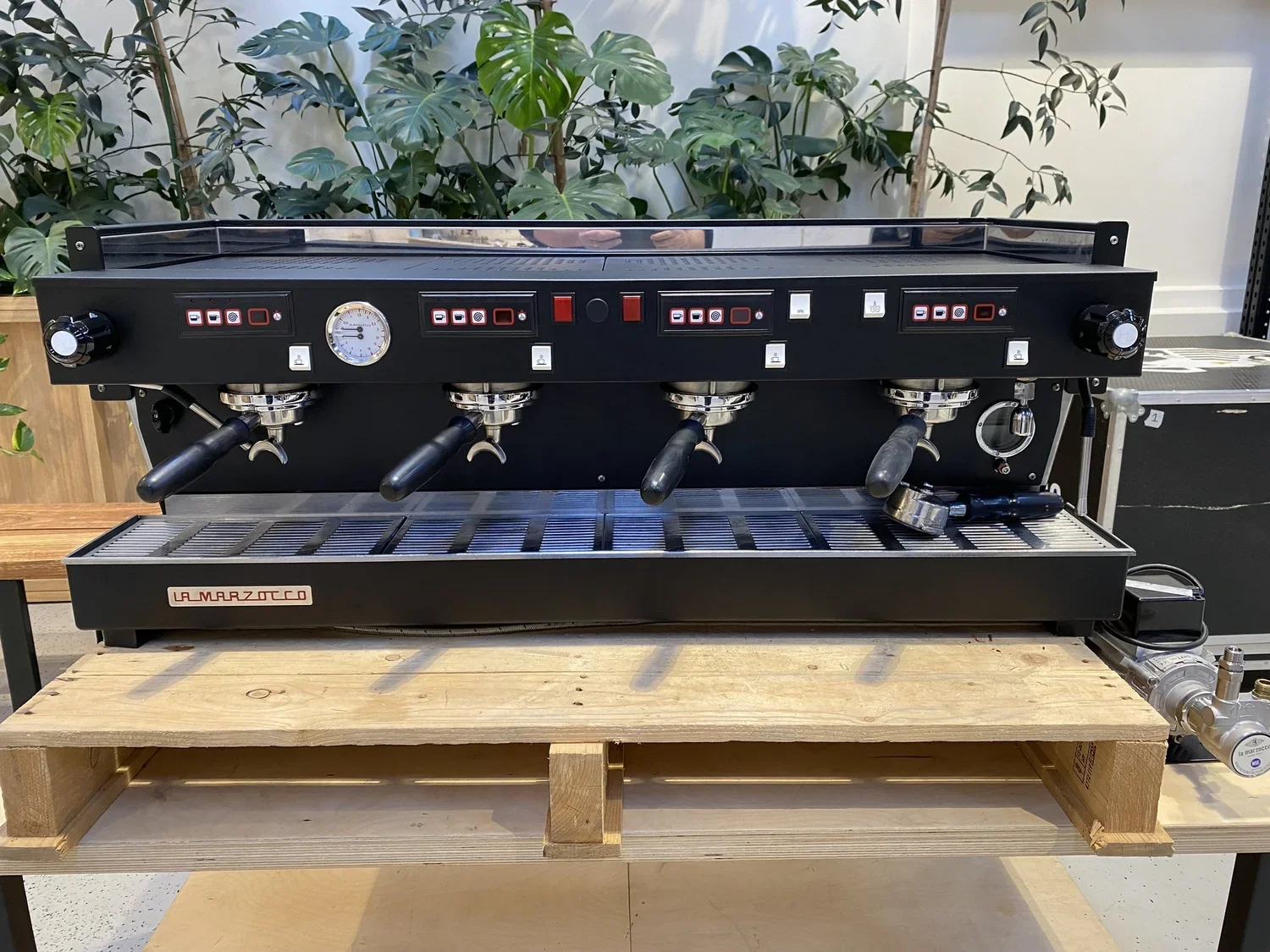 A black La Marzocco espresso machine with four group heads, placed on a wooden table in front of green plants.