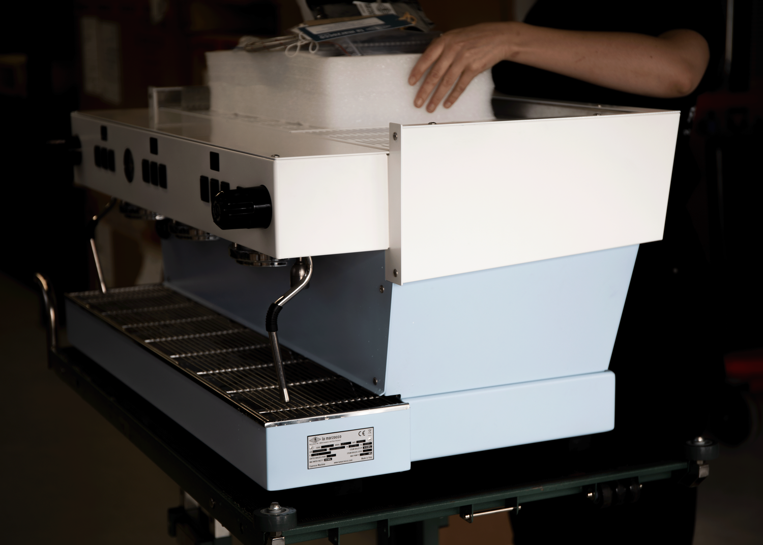 A person loading items into the top of a white and light blue espresso machine with a metal drip tray and black control knob.