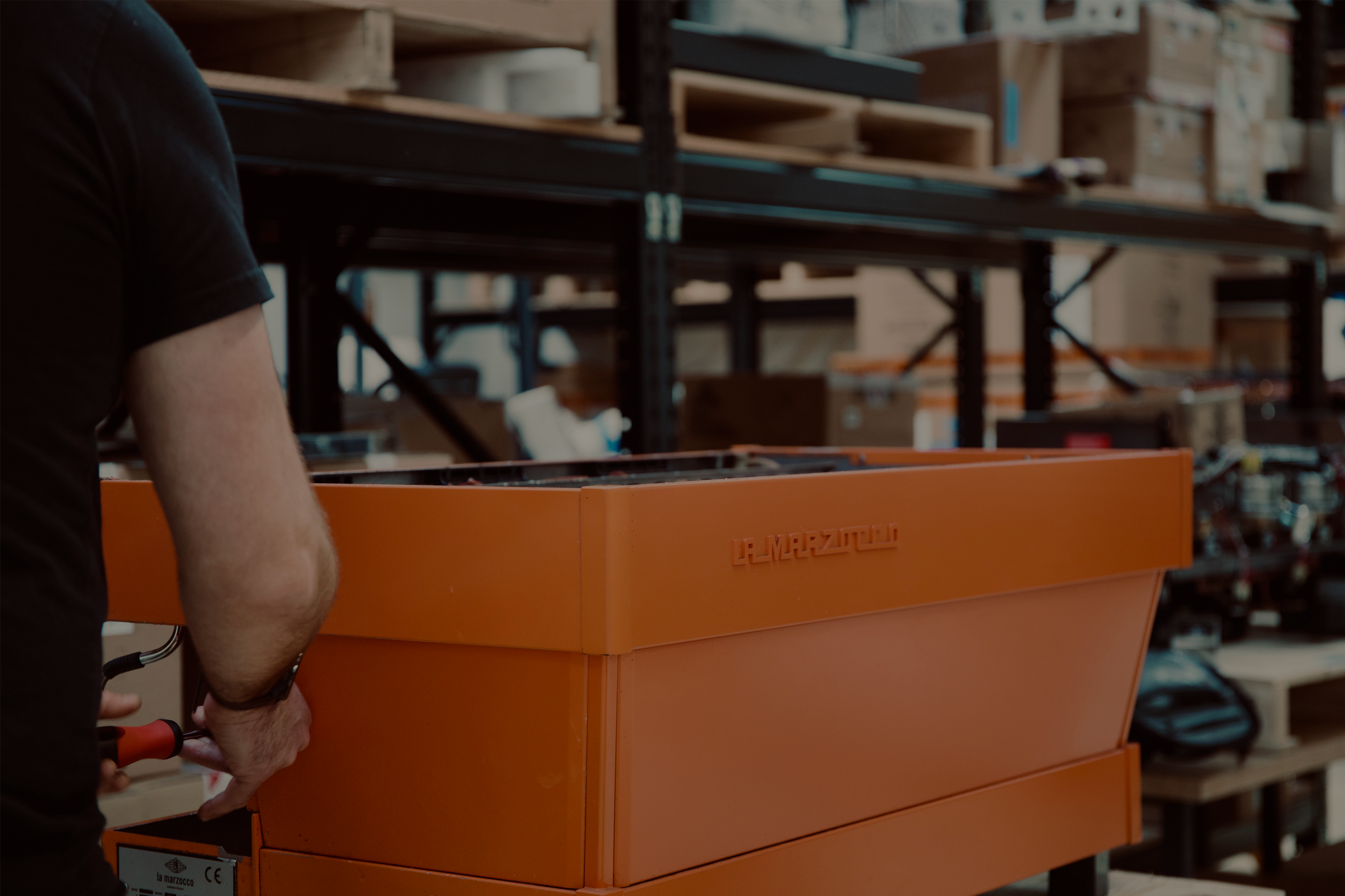 A Brewtech technician working with an orange La Marzocco tool chest in a warehouse or workshop surrounded by shelves with boxes and various equipment.