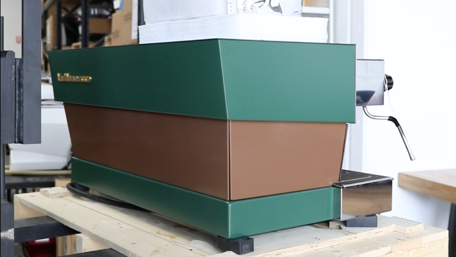 A vintage green and brown espresso machine with a lever on the right side, placed on a wooden table in a workshop or storage area.