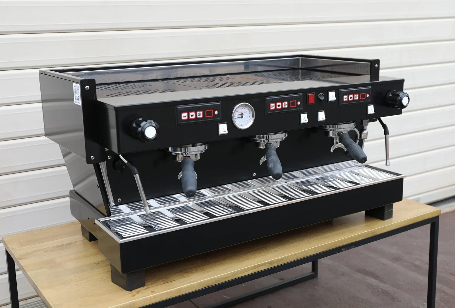 Black commercial espresso machine with three group heads on a wooden table against a beige wall.