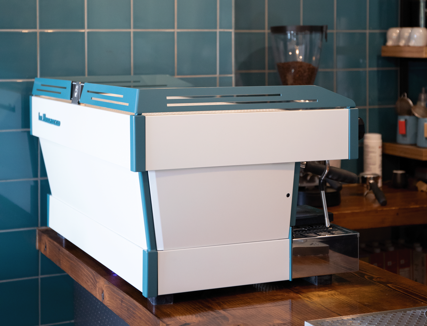 A professional espresso machine on a wooden countertop in a cafe setting, with a teal tiled wall in the background.