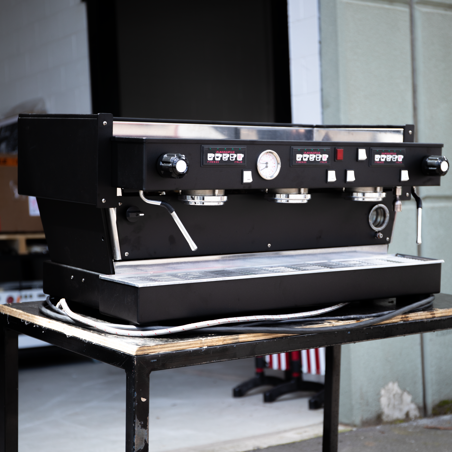 Black commercial espresso machine on a wooden table outdoors.