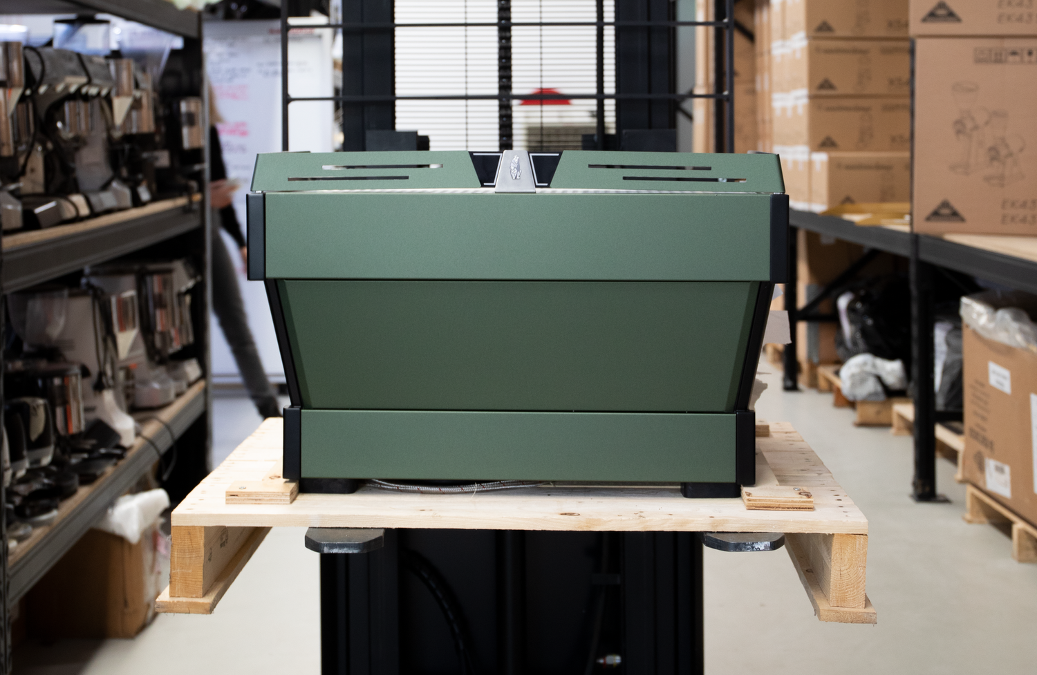 A green industrial machine on a wooden pallet in a warehouse with shelves of products and boxes.