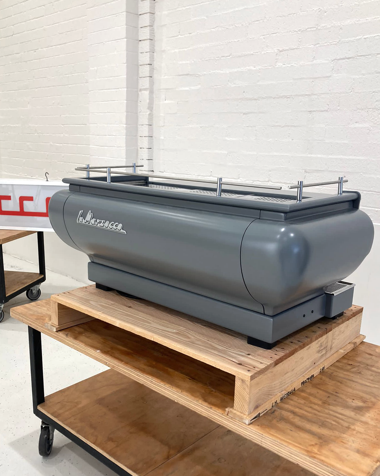 A large gray display case with the brand name La Marzocco on it, placed on a wooden table in front of a white brick wall.