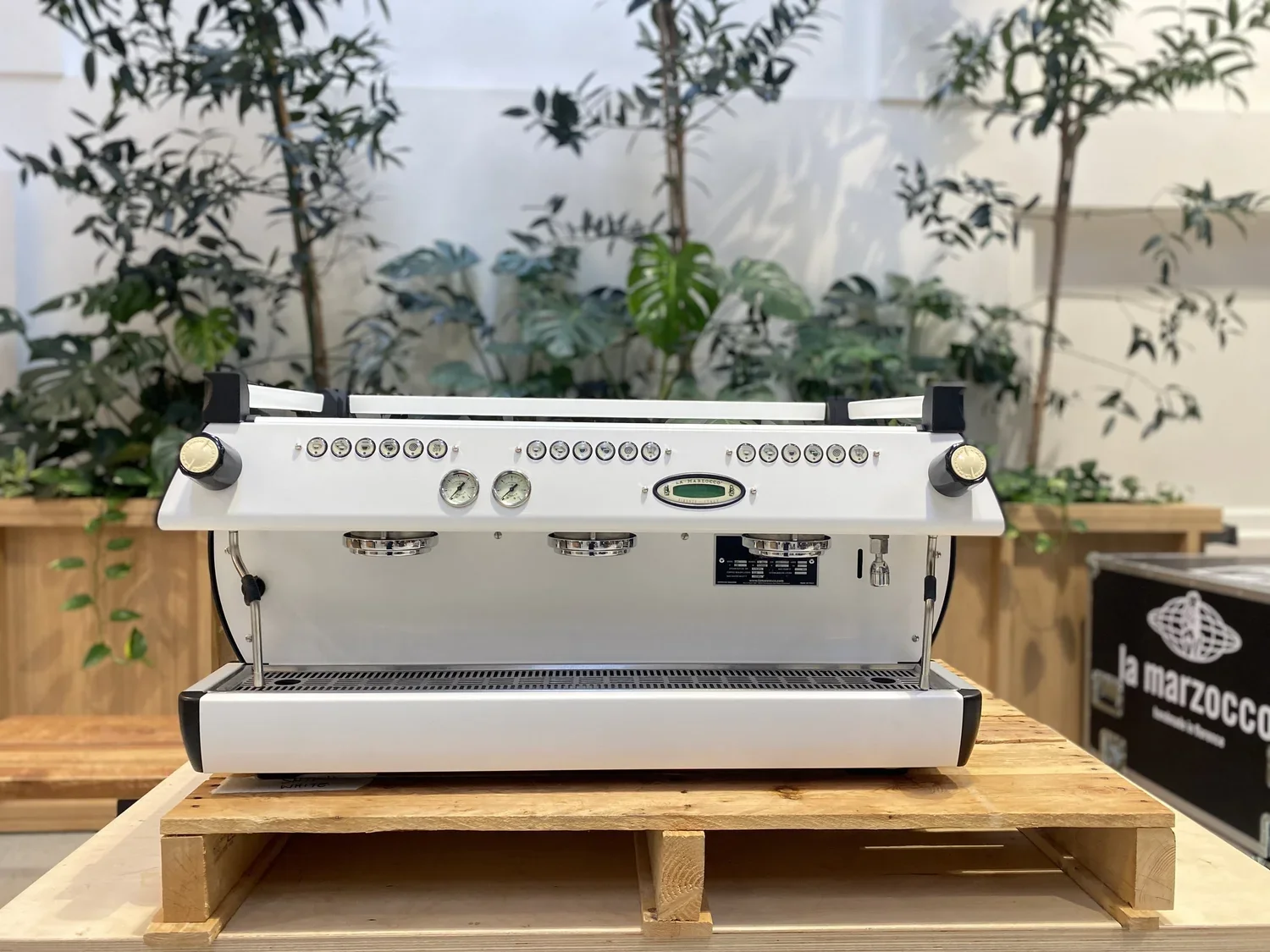 White commercial espresso machine with multiple gauges, buttons, and dials on a wooden table with plants in the background.