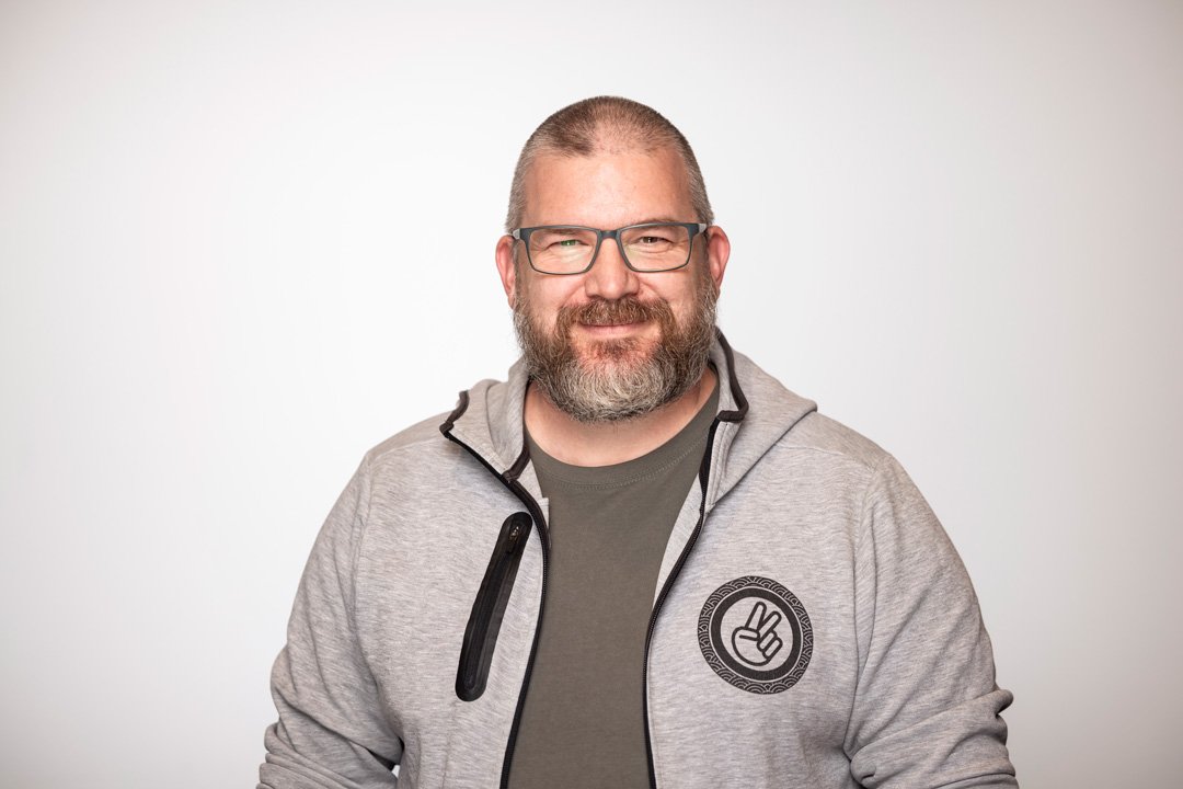 Neil, CEO and principal consultant at Searchlight, experienced Critical Network Infrastructure, a smiling man with glasses, a beard, and short hair wearing a gray hoodie with a peace sign patch, standing against a plain background.