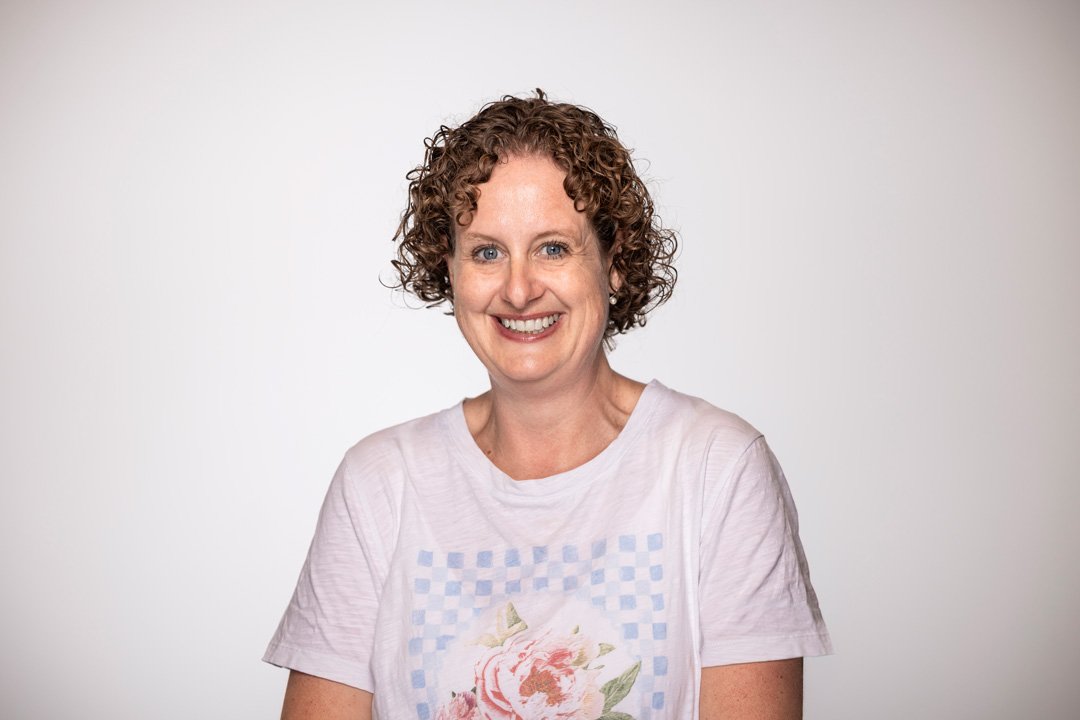 Rebekah, finance administrator at Searchlight - Enterprise network solutions NZ,  woman with curly brown hair smiling at the camera against a plain white background, wearing a white T-shirt with a floral design.