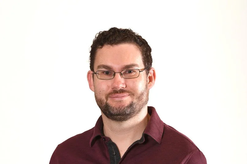 Shane, Senior Software Developer at Searchlight, Resilient network architecture Auckland New Zealand, man with curly hair, glasses, and a beard, wearing a maroon shirt, against a white background.