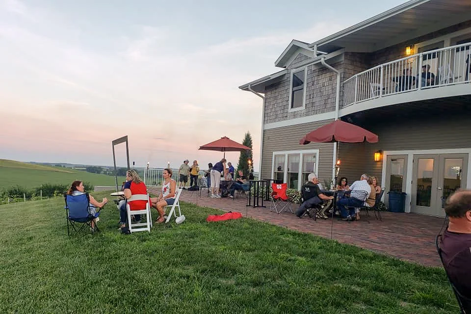 People gathering and socializing on a grassy lawn and patio area next to a large house with a balcony, during sunset.
