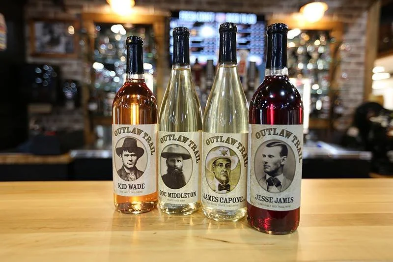 Four bottles of wine with labels featuring portraits of historical outlaws Kid Wade, Doc Middleton, James Capone, and Jesse James, lined up on a wooden bar in a dimly lit bar or restaurant.
