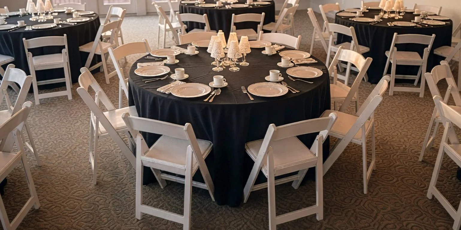 Round banquet table with black tablecloth, set with white plates, cups, saucers, silverware, and small lamps with shades, surrounded by white chairs in a decorated event space.