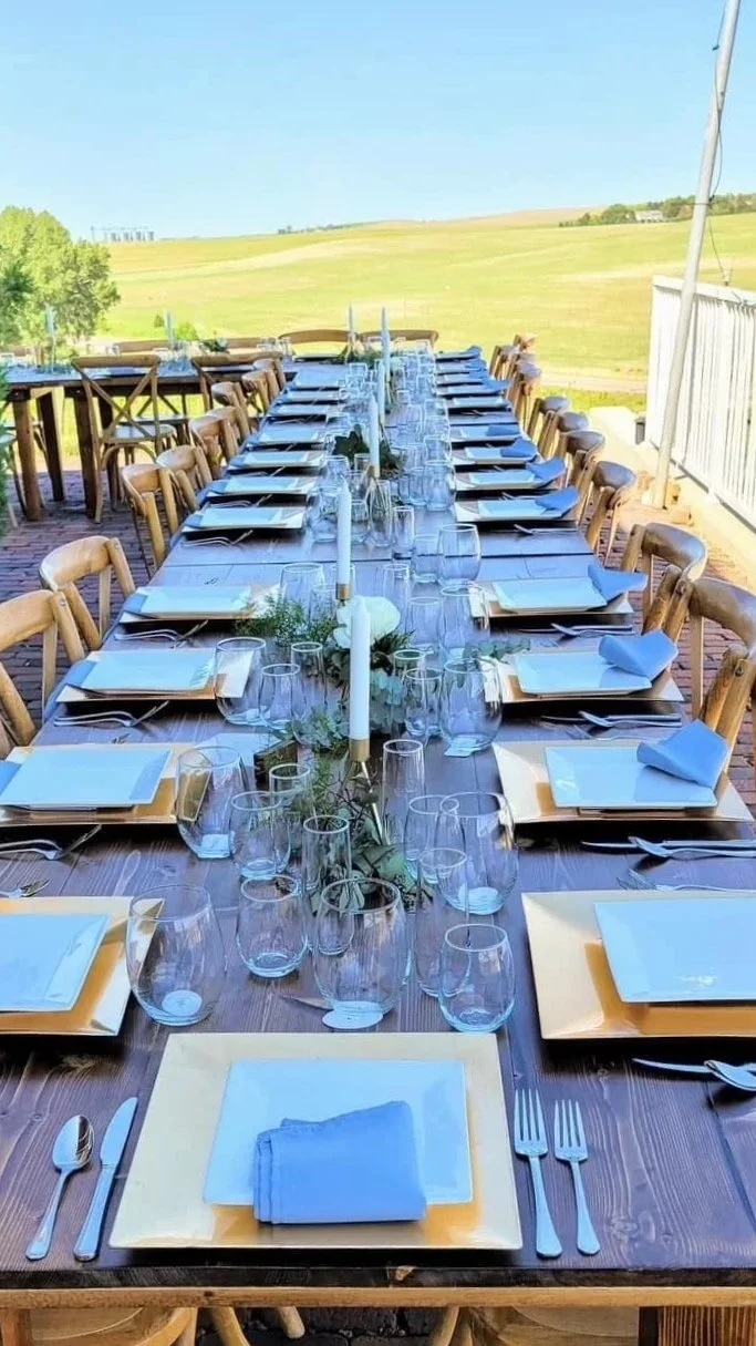 Long outdoor dining table set with napkins, glasses, and candle centerpieces, overlooking a green landscape.