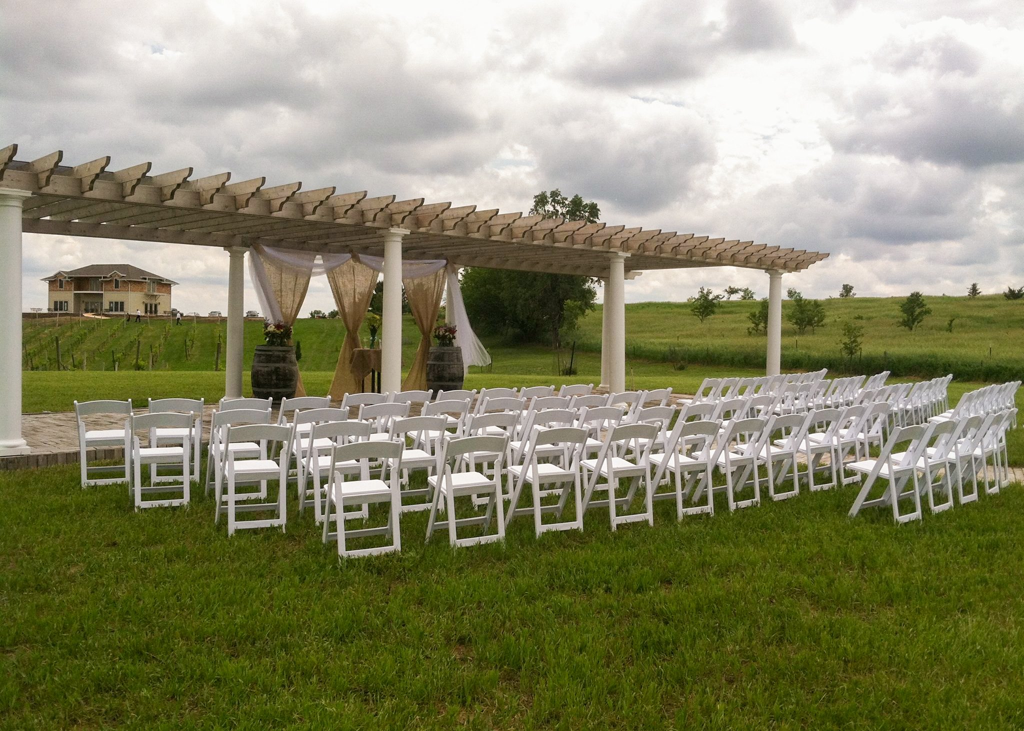 Outdoor wedding setup with white chairs arranged in rows under a wooden pergola with curtains and flower decorations, on a green lawn with a building and cloudy sky in the background.