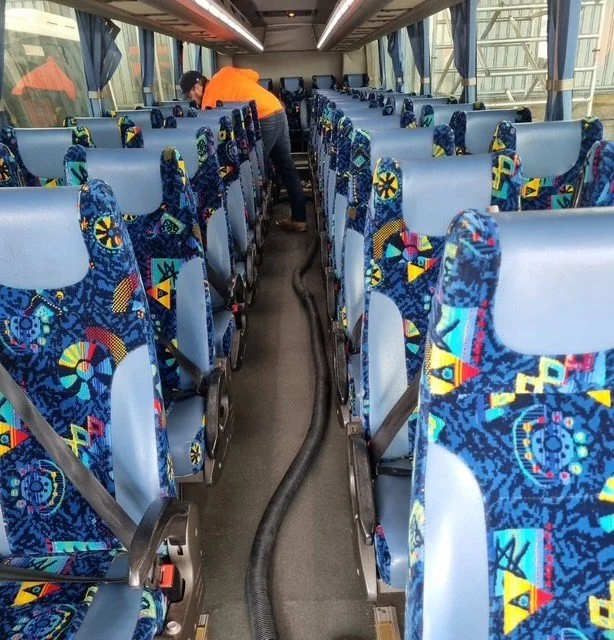 Inside a bus with rows of colorful patterned seats on both sides, and a person in an orange shirt cleaning or inspecting the aisle.