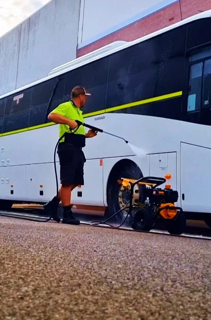 A person in a bright green shirt and shorts is cleaning the wheel of a large bus using a pressure washer. The bus is parked on a concrete surface outdoors, with a building visible in the background, and a pressure washer machine is on the ground nearby.