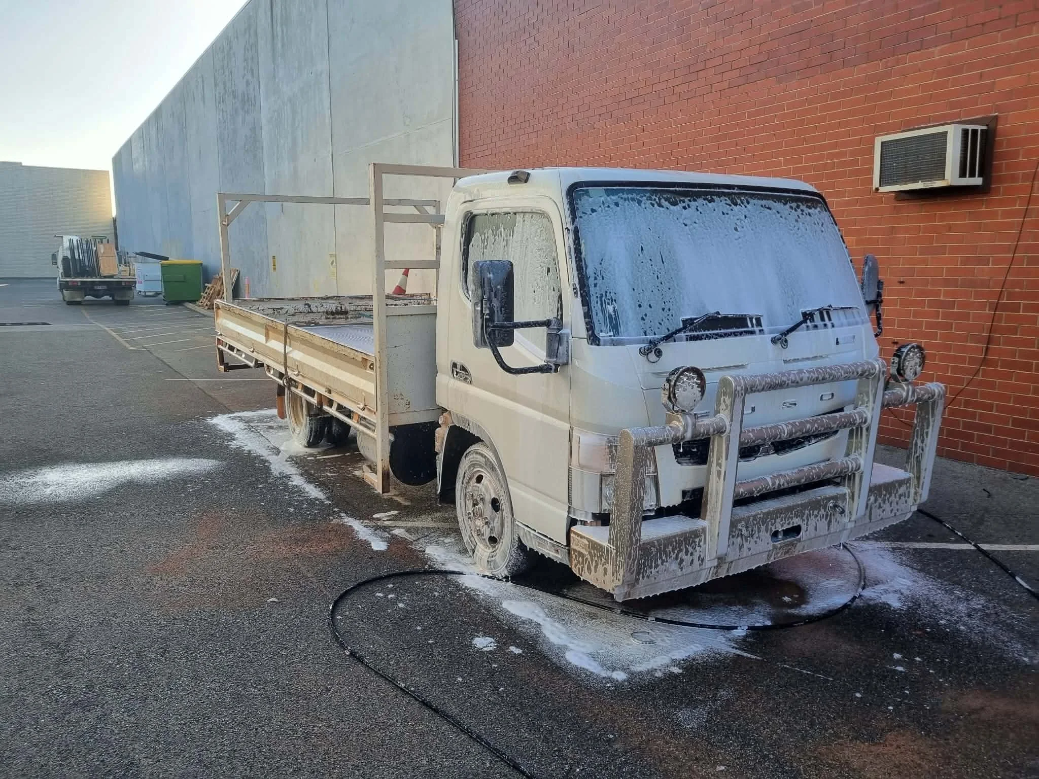 A small white flatbed truck washing with soap and water in an alleyway next to a brick wall, with an air conditioning unit on the wall.