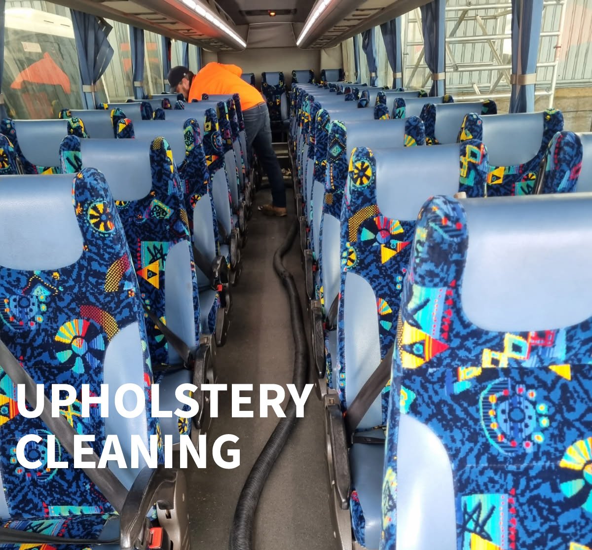 Inside a bus with patterned blue seats, a worker in an orange shirt is cleaning the upholstery with cleaning equipment, including a vacuum hose. The bus has windows on both sides with blue curtains.