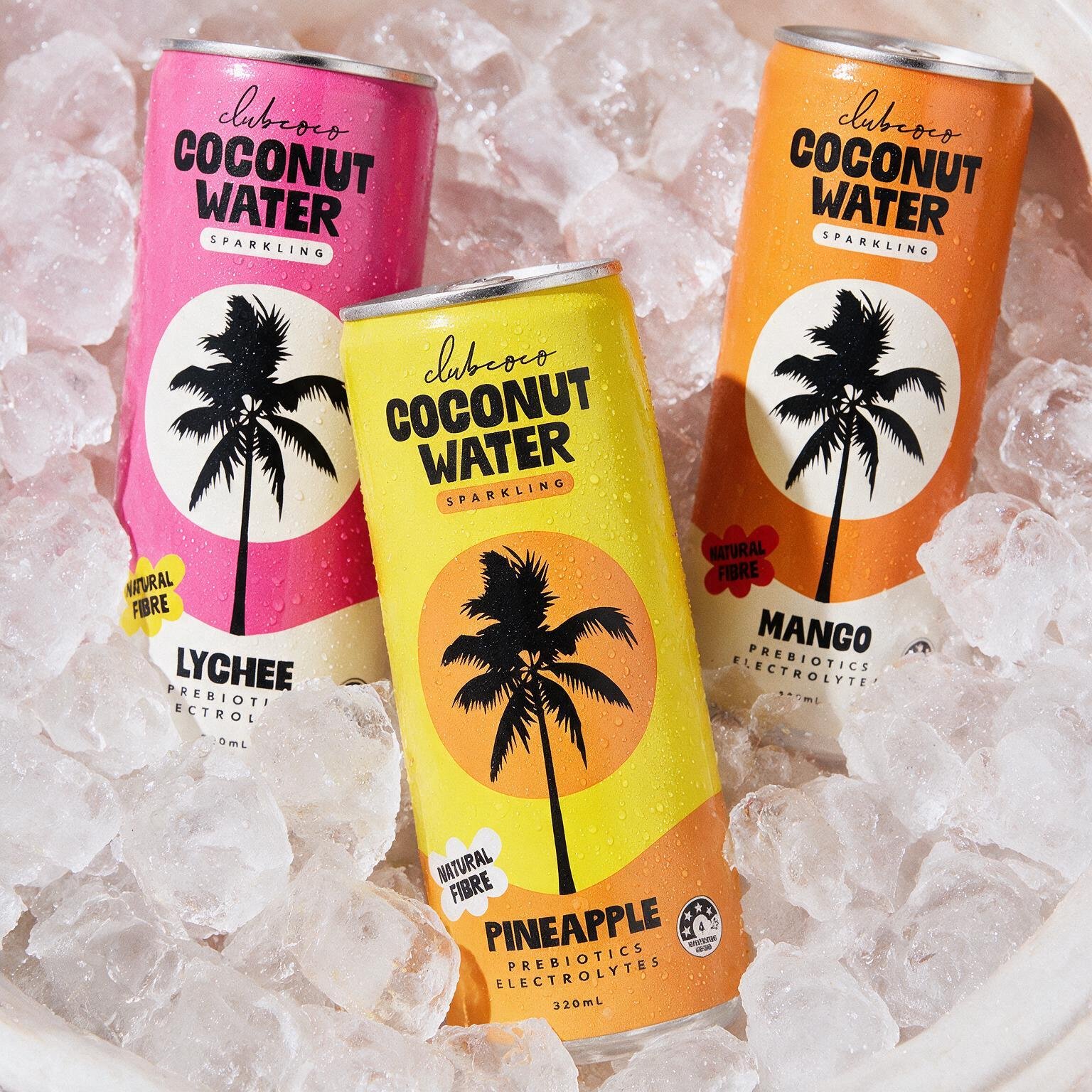 Three cans of Club Coco sparkling coconut water with colorful designs, placed on ice. The cans are pink, yellow, and orange, each featuring a black palm tree silhouette and labeled with flavor: lychee, pineapple, and mango.