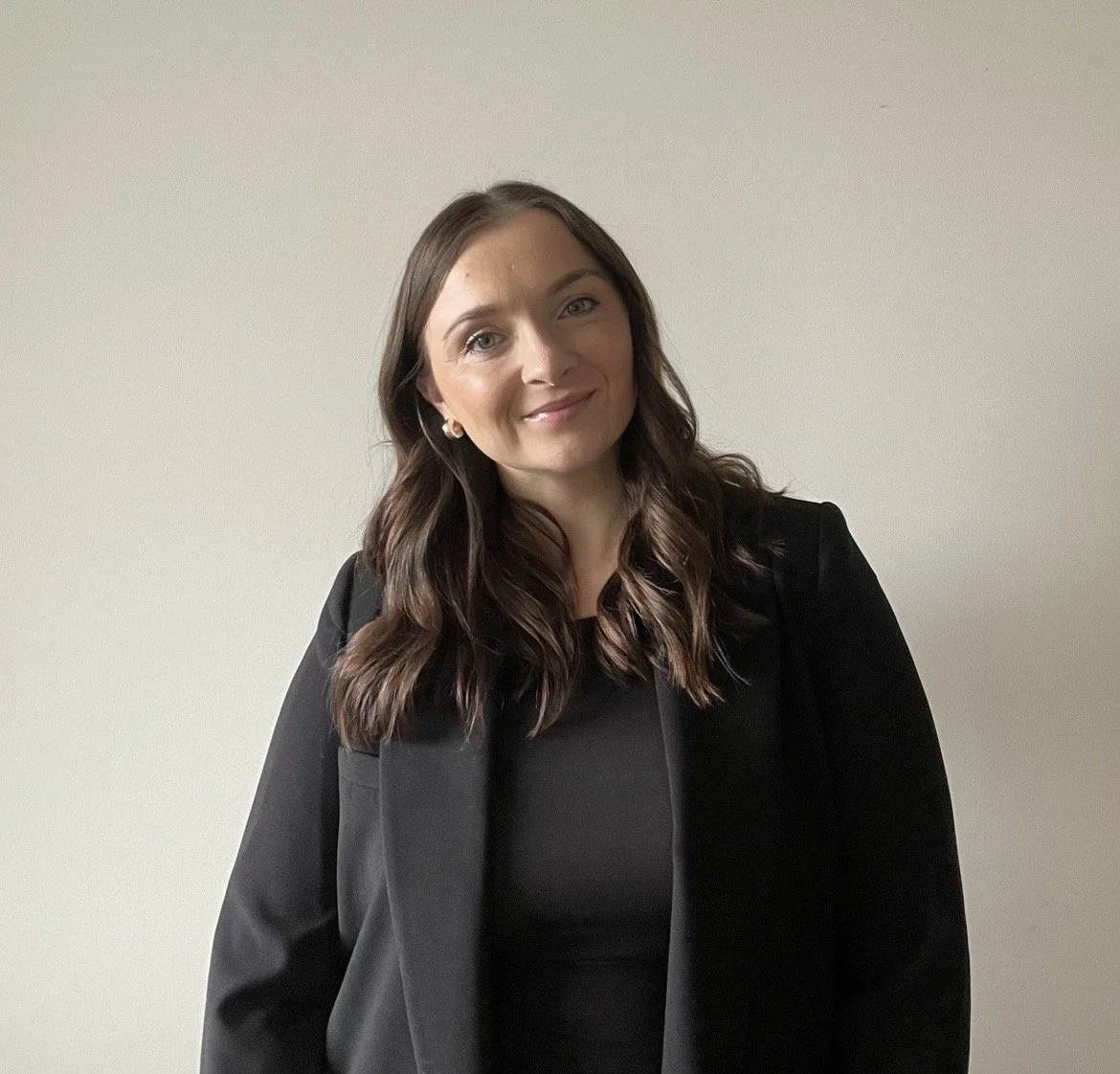 A woman with long brown hair, wearing a black blazer and black top, smiling and standing against a plain light-colored wall.
