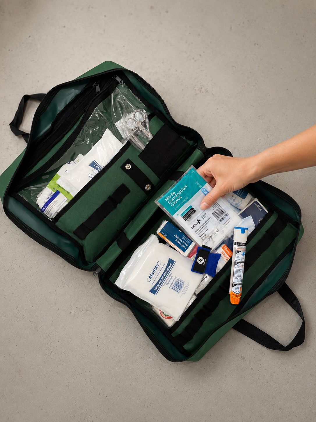 Open first aid kit with medical supplies, including gauze, scissors, ointment, and gloves, on a light-colored surface, with a hand placing a pack of nitrile examination gloves inside.