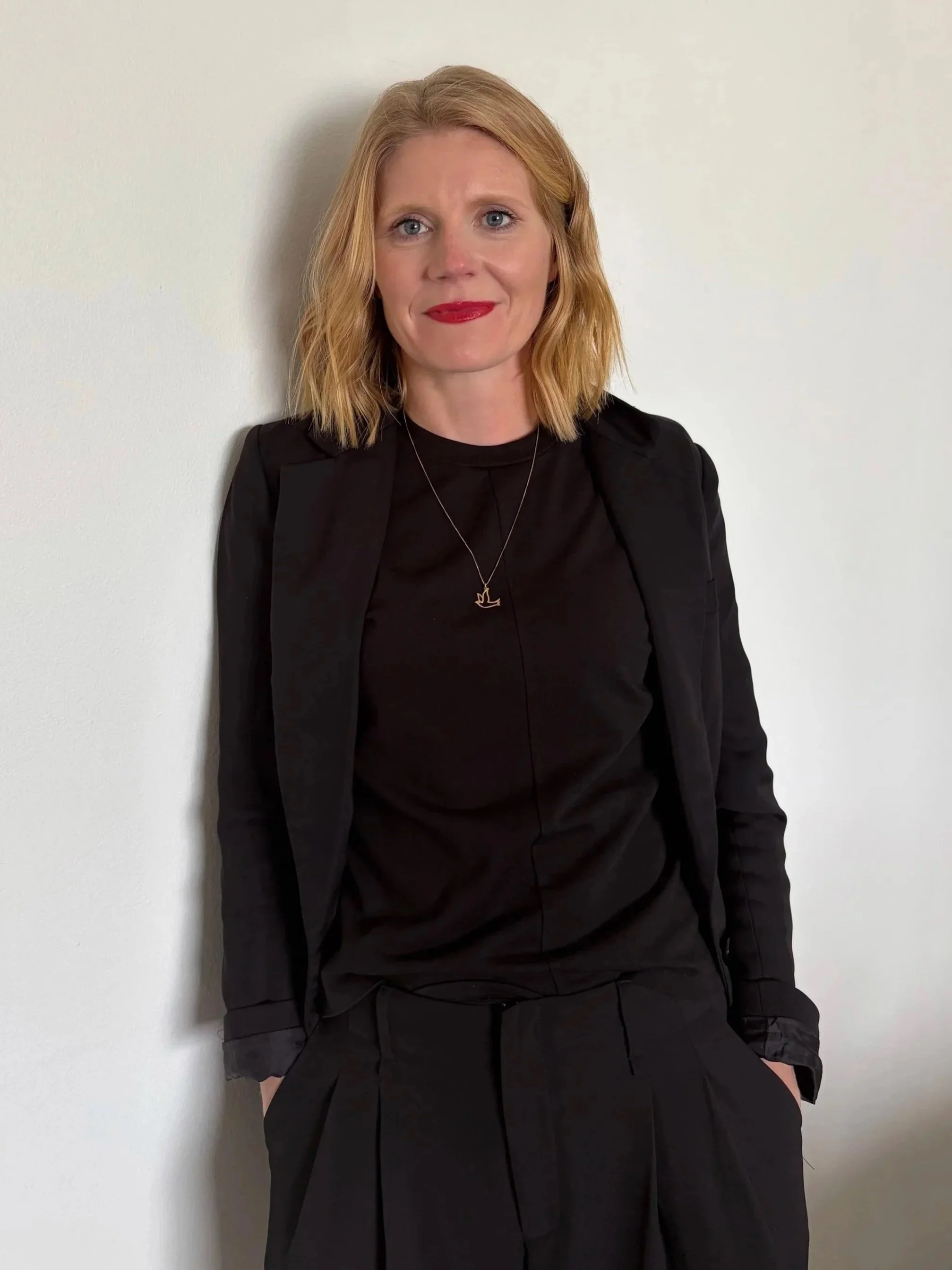 Portrait of a woman with blonde hair and red lipstick, wearing a black blazer and black dress, sitting against a plain white wall.