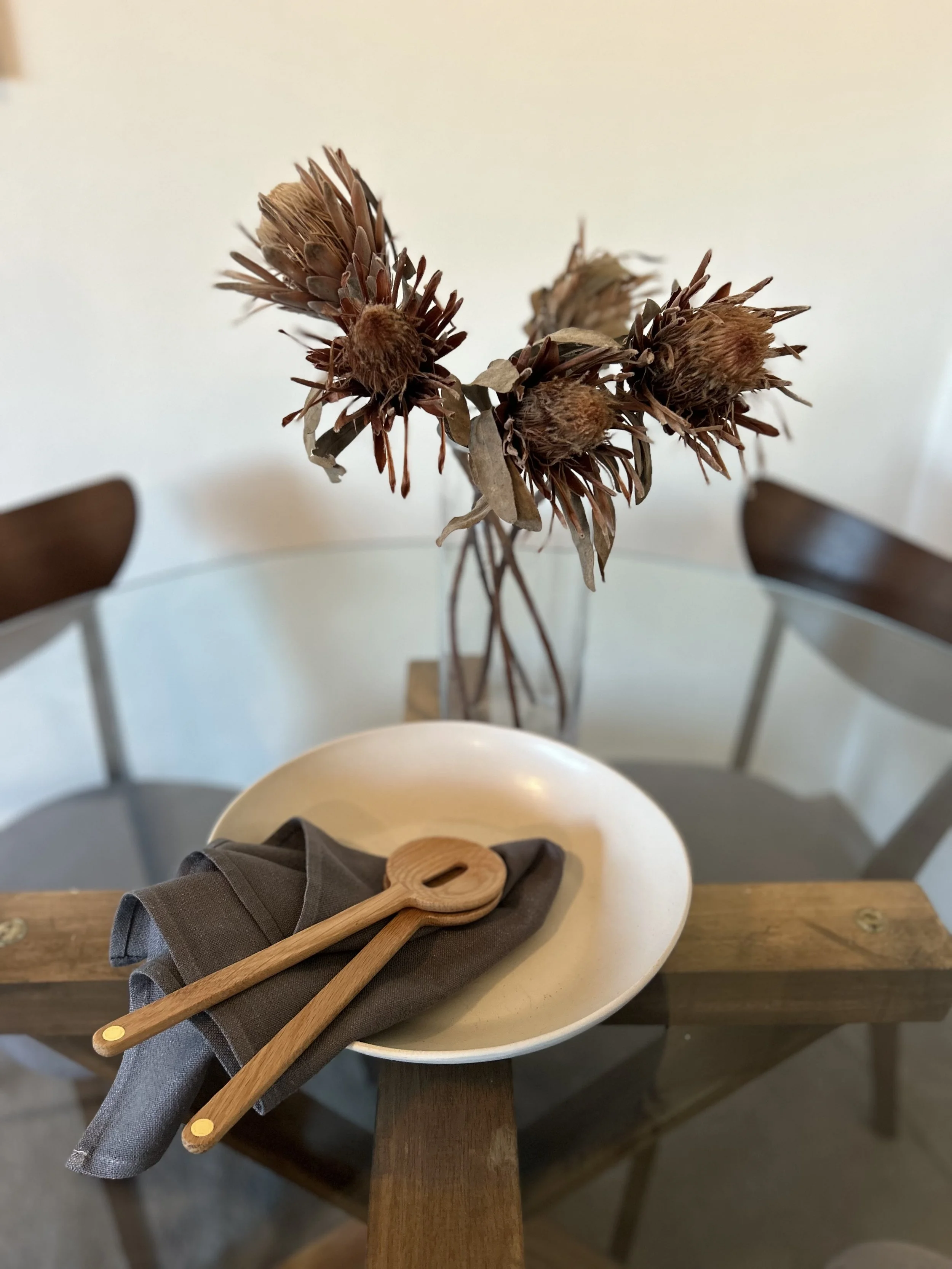 Dried flowers arranged in a glass vase on a wooden dining table. Two wooden serving utensils are resting on a white plate with a grey napkin, all placed on the table. In the background, there are two chairs.