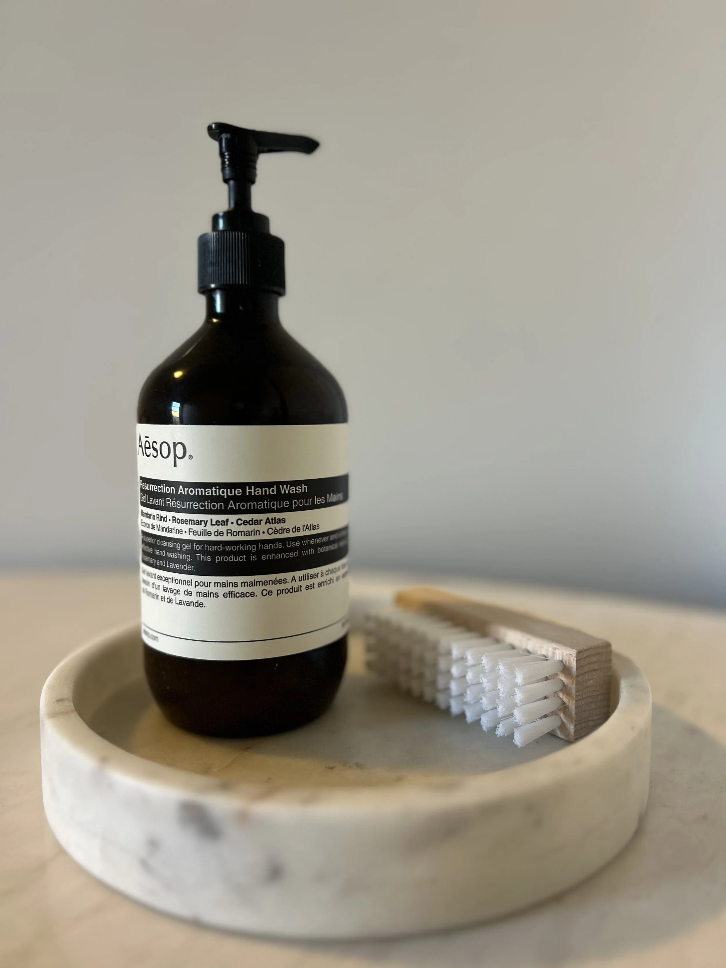A dark brown bottle of Aesop Resurrection Aromatique Hand Wash placed on a round marble soap dish, with white toothbrushes on a wooden holder in the background, on a beige surface.