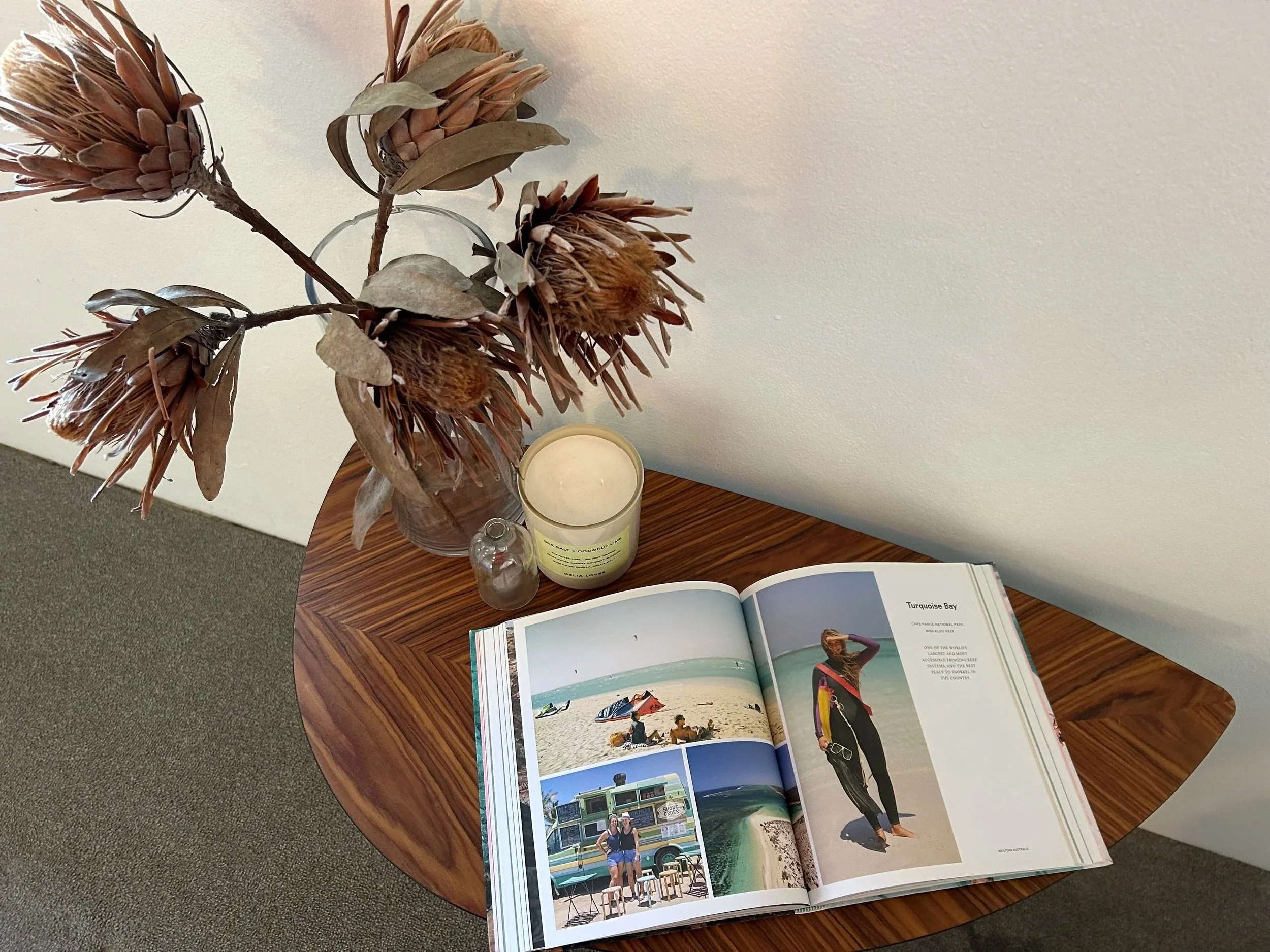 A wooden table with a magazine opened to pages featuring beach and surfing images, a glass vase with dried protea flowers, a small clear glass bottle, a lit white candle, and an open book or booklet.