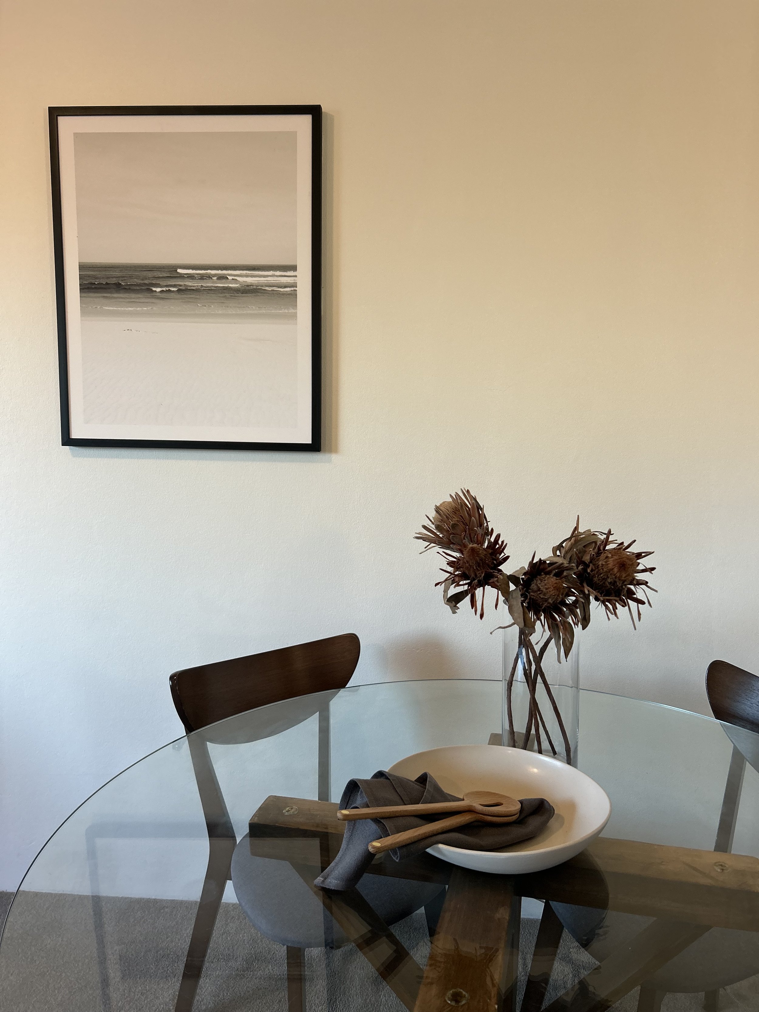 A round glass dining table with a brown wooden base, four chairs with dark brown wooden backs, and a white high chair with drying wood spoons and a dark cloth. A vase with dried flowers is on the table. A wall with a framed black and white beach scene photograph is in the background.