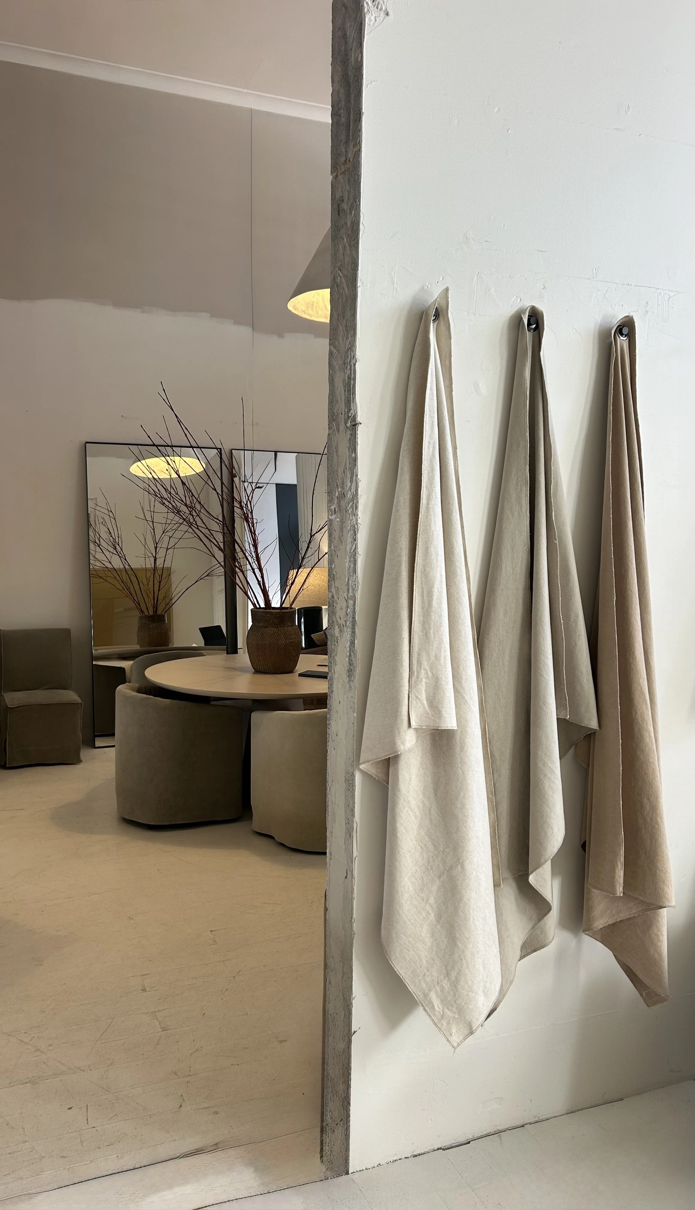 Interior view of a room divided by a partially exposed wall. On the right, beige and cream-colored towels hang on hooks. On the left, there is a round table with a vase containing bare branches, a mirror, and some chairs.